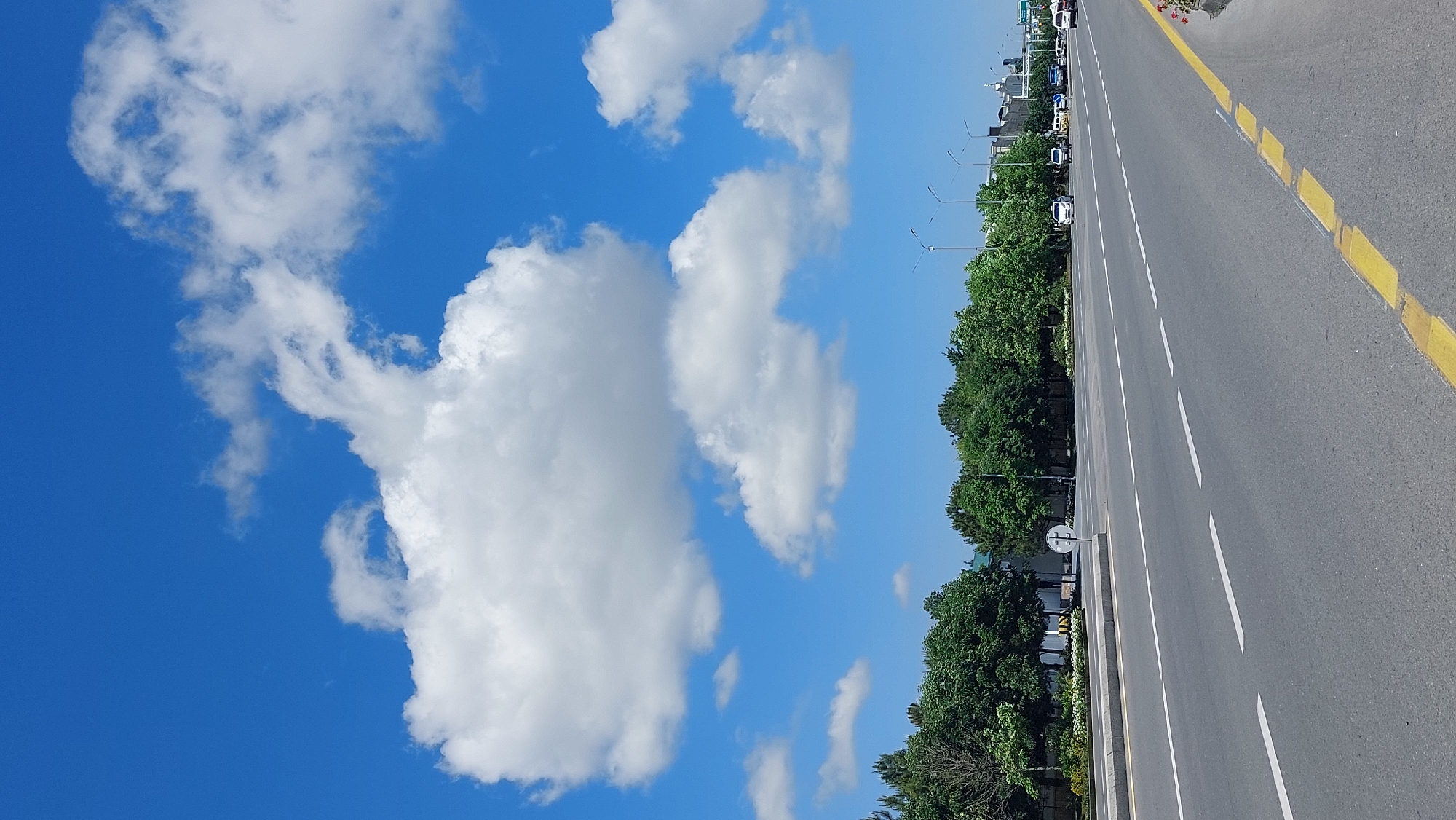The image depicts a section of road in Samarkand City, Uzbekistan. The sky, predominantly blue, is populated with scattered white clouds. A road occupies the right side of the frame, marked with white lines delineating lanes and a yellow line along the shoulder. Green trees line the edge of the road. Street lights are visible in the distance along the roadside. Vehicles are parked along the side of the road in the distance.