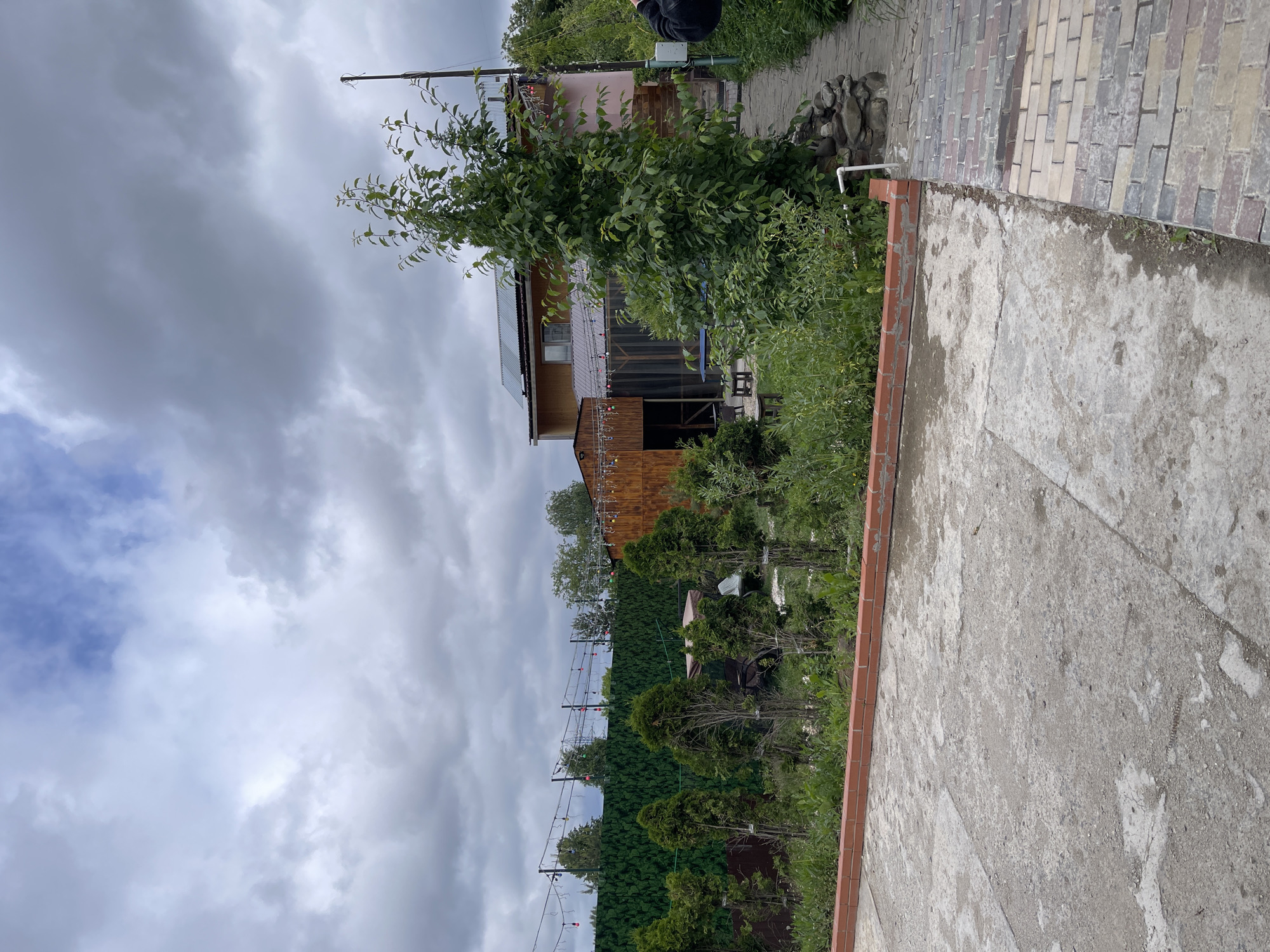 The image shows a residential area in Kommunarka, Russia, under a cloudy sky. A two-story wooden house is visible behind green foliage and trees. String lights are hung between the trees. The foreground features a concrete patio bordered with brick. A walkway made of multi-colored paving stones is also present. A water pipe protrudes from the ground near the walkway. A person is partially visible in the upper right corner. The environment appears to be a private garden or backyard.