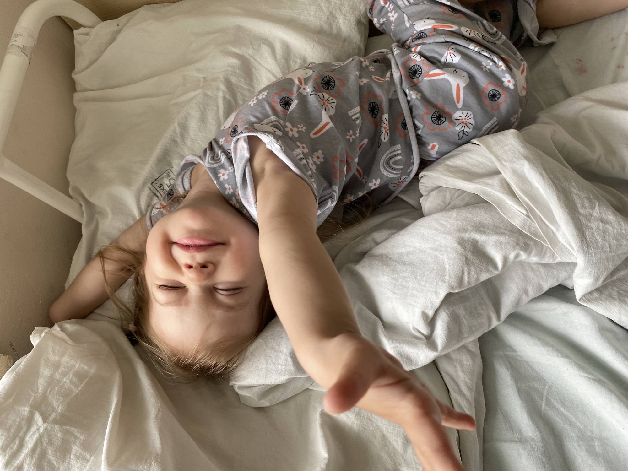 A young girl lies on a bed covered with white sheets and blankets. She is wearing grey pajamas with a rabbit and floral pattern. Her eyes are closed and she is smiling slightly. Her right arm is extended towards the camera. The bed has a white metal frame. The location is unknown.