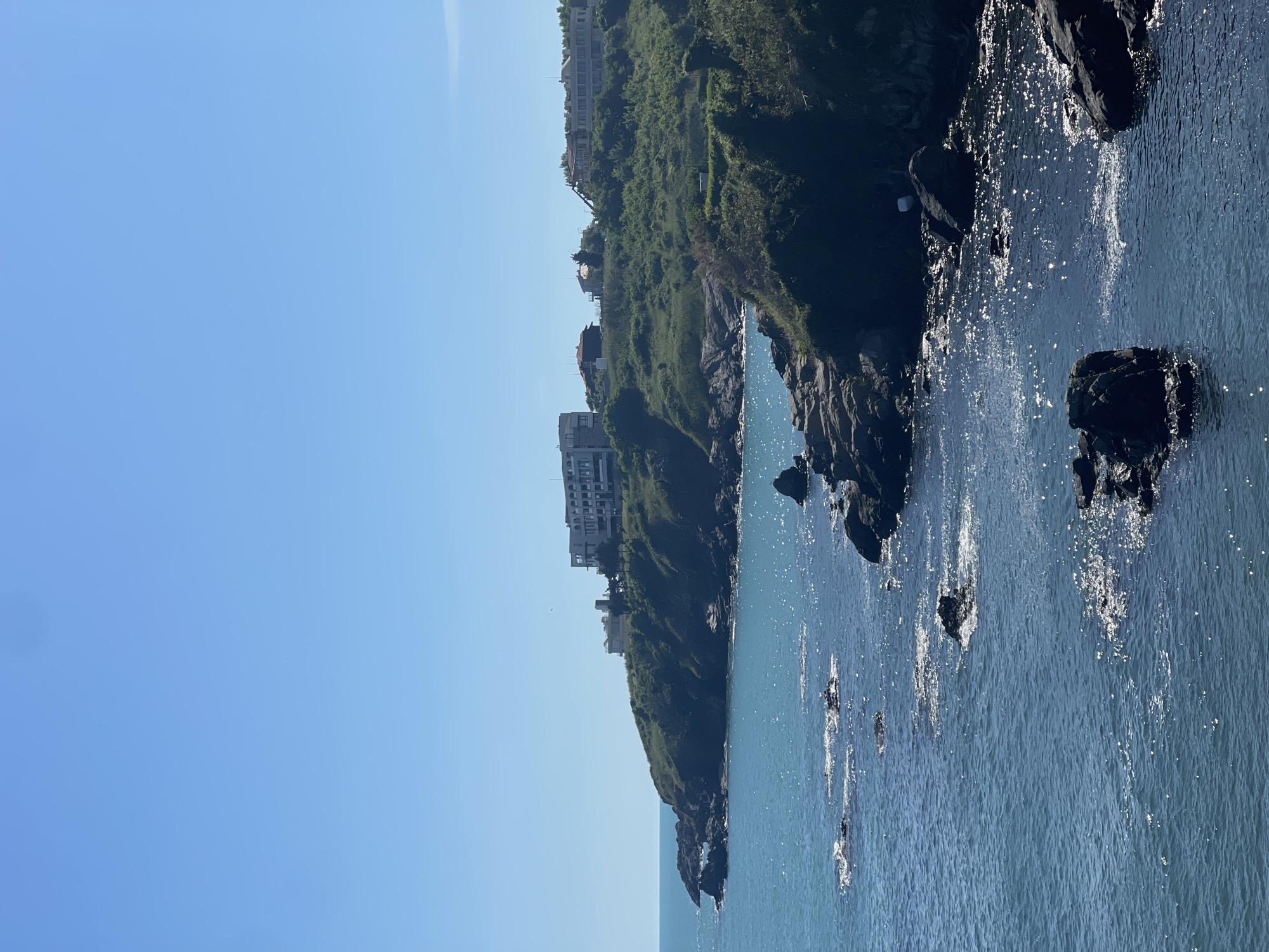 The image shows a coastline view in Sozopol, Bulgaria. Buildings are visible on top of a cliff covered with greenery. Below the cliff, the ocean meets the rocky shoreline. Sunlight reflects on the water's surface, creating a shimmering effect. The sky is clear and blue.