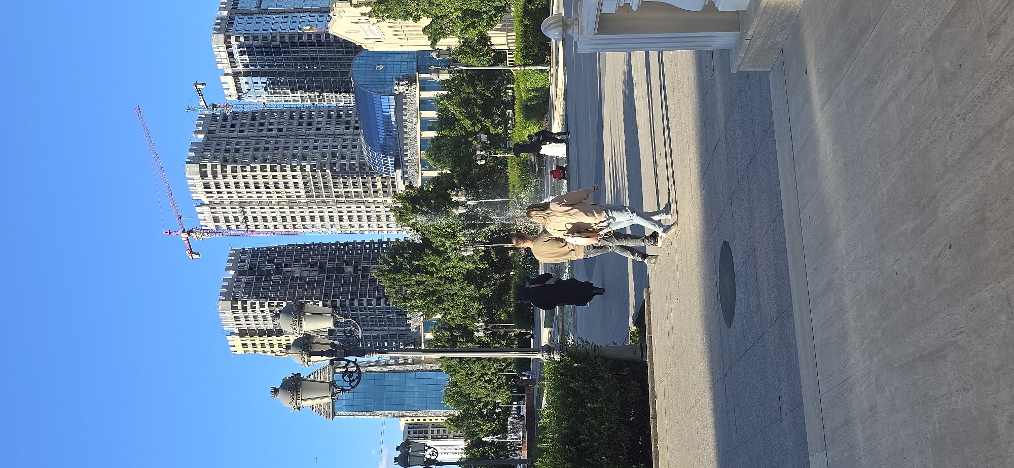 Here is a description of the image:
The image captures a street scene in what appears to be a modern urban environment. Tall buildings, some under construction with cranes visible, rise in the background, suggesting a city undergoing development. The sky is clear and blue.
In the foreground, a couple is walking along a pedestrian walkway. The man is wearing a tan jacket and dark pants, while the woman is wearing a similar jacket and light jeans. There are other people and trees lining the walkway. There are also period-style streetlights along the walkway.
The image does not state that this is Grozny, Russia.