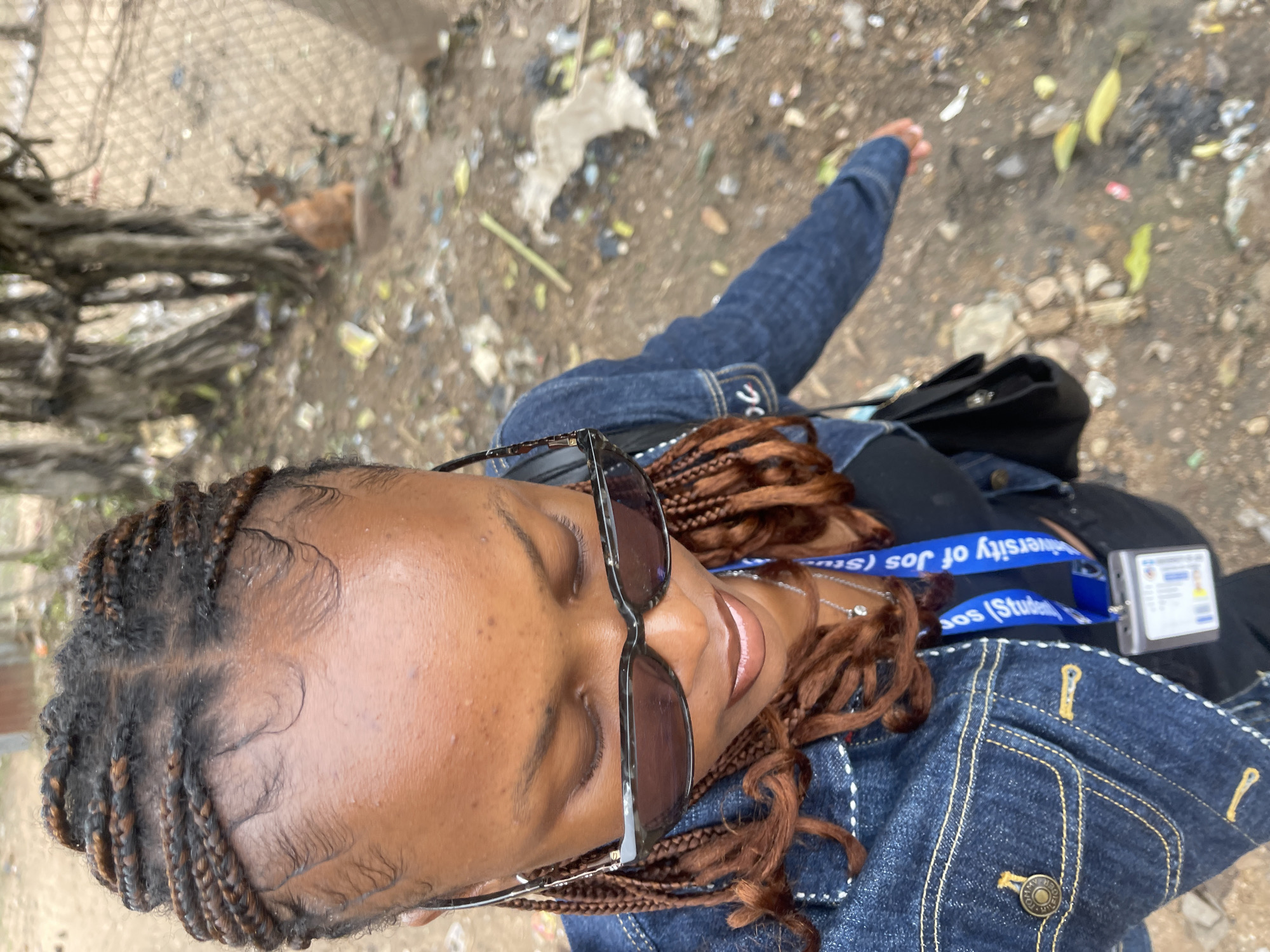The image shows a young woman with braided hair and sunglasses, wearing a denim jacket and a black top. She is standing outdoors in what appears to be a street with debris on the ground. In the background, there are trees and a chain-link fence. A cat can be seen in the background. The woman is wearing a lanyard with an ID card. There is no specific information in the image to identify the location as Rafin Par, Nigeria.