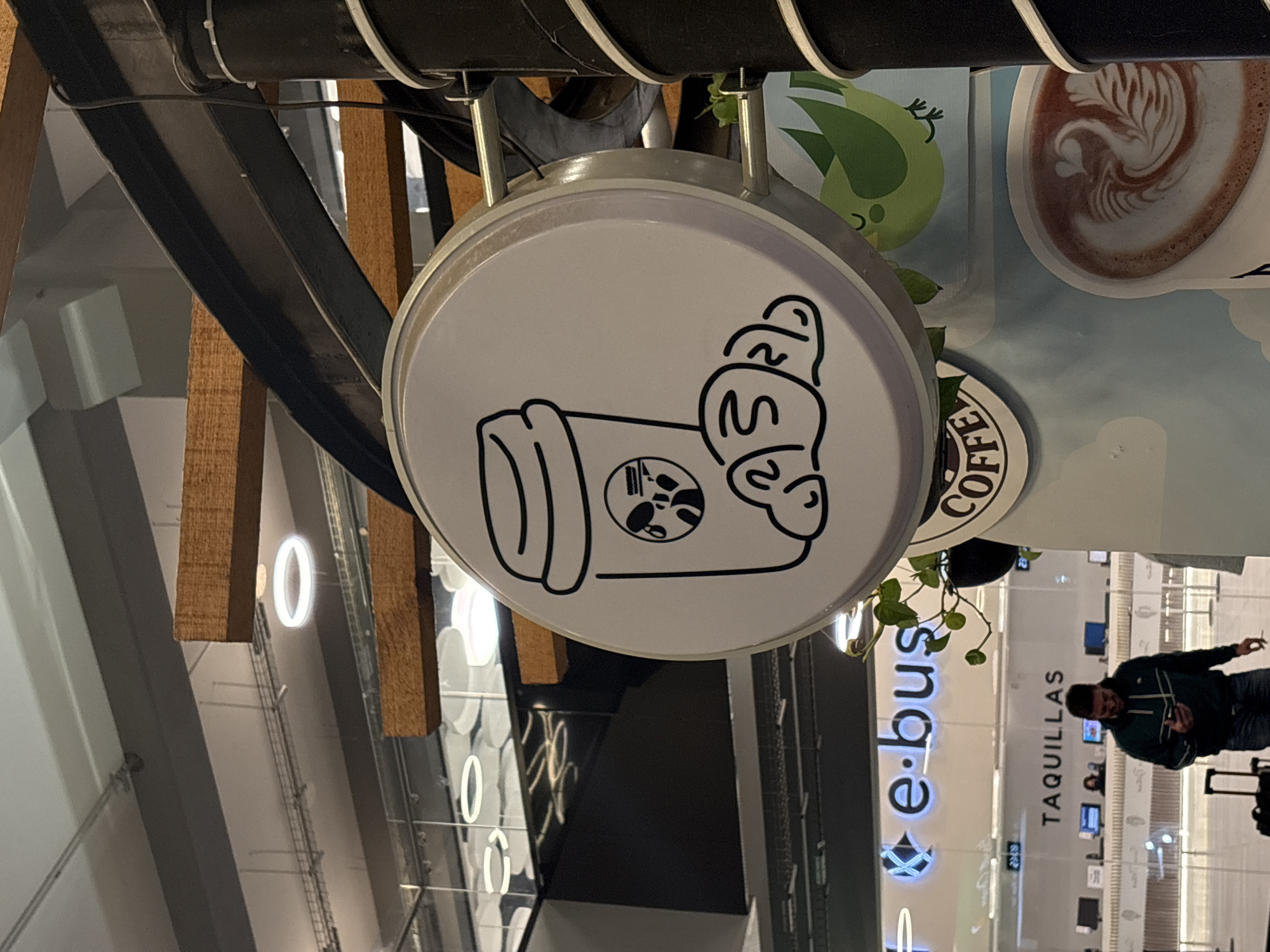 The image shows a coffee shop sign in Puebla, Mexico. The sign is round, white, and features a stylized drawing of a coffee cup with whipped cream. The sign hangs from a black, metal structure. In the background, a man wearing a dark jacket is visible, standing in front of a counter labeled "e-bus TAQUILLAS." The location appears to be an indoor transportation hub or terminal.