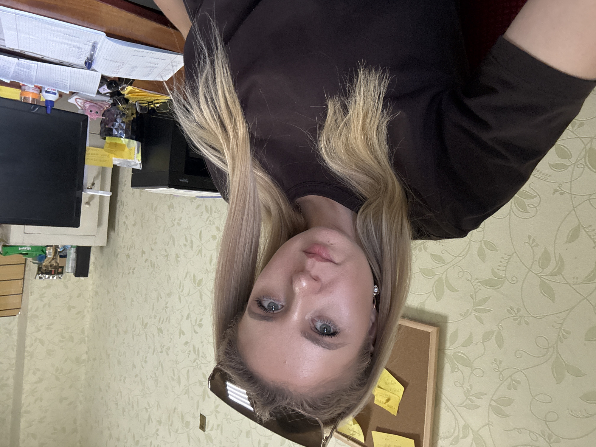 A young woman with blonde hair and blue eyes is pictured from the chest up, looking directly at the camera. She is wearing a dark brown t-shirt and has sunglasses perched on her head. Her left ear is adorned with a silver hoop earring. Behind her, a wall is covered in light green floral wallpaper. To her left, a bulletin board is partially visible, featuring several yellow sticky notes. To her right, a desk is set up with various items including a printer, a computer monitor, notebooks, pens, a bottle of glue, and a small pink toy. The overall environment appears to be an indoor office or workspace.