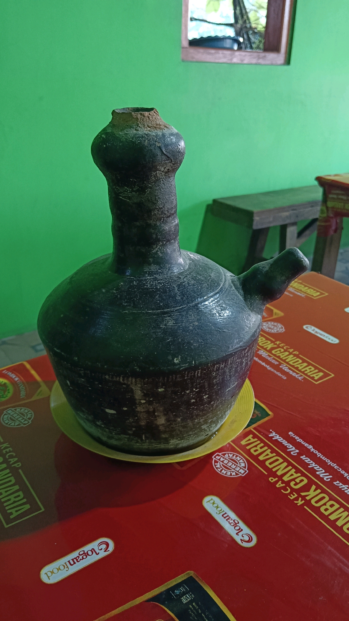 An antique, dark-colored ceramic jug with a narrow neck and a spout sits on a yellow ceramic saucer. The jug is positioned on a red surface with some text visible, including "KECAP GANDARIA" and "Loganfood." In the background, a bright green wall is visible, with a wooden stool and a glimpse of a window with reflections of trees and sky. The scene appears to be indoors, possibly in a home or shop in Jogorogo, Indonesia, given the context. The lighting is natural and appears to be from the side, casting shadows on the jug. There are no people or animals visible in the image. The overall impression is of a still life, highlighting the texture and form of the ceramic jug.