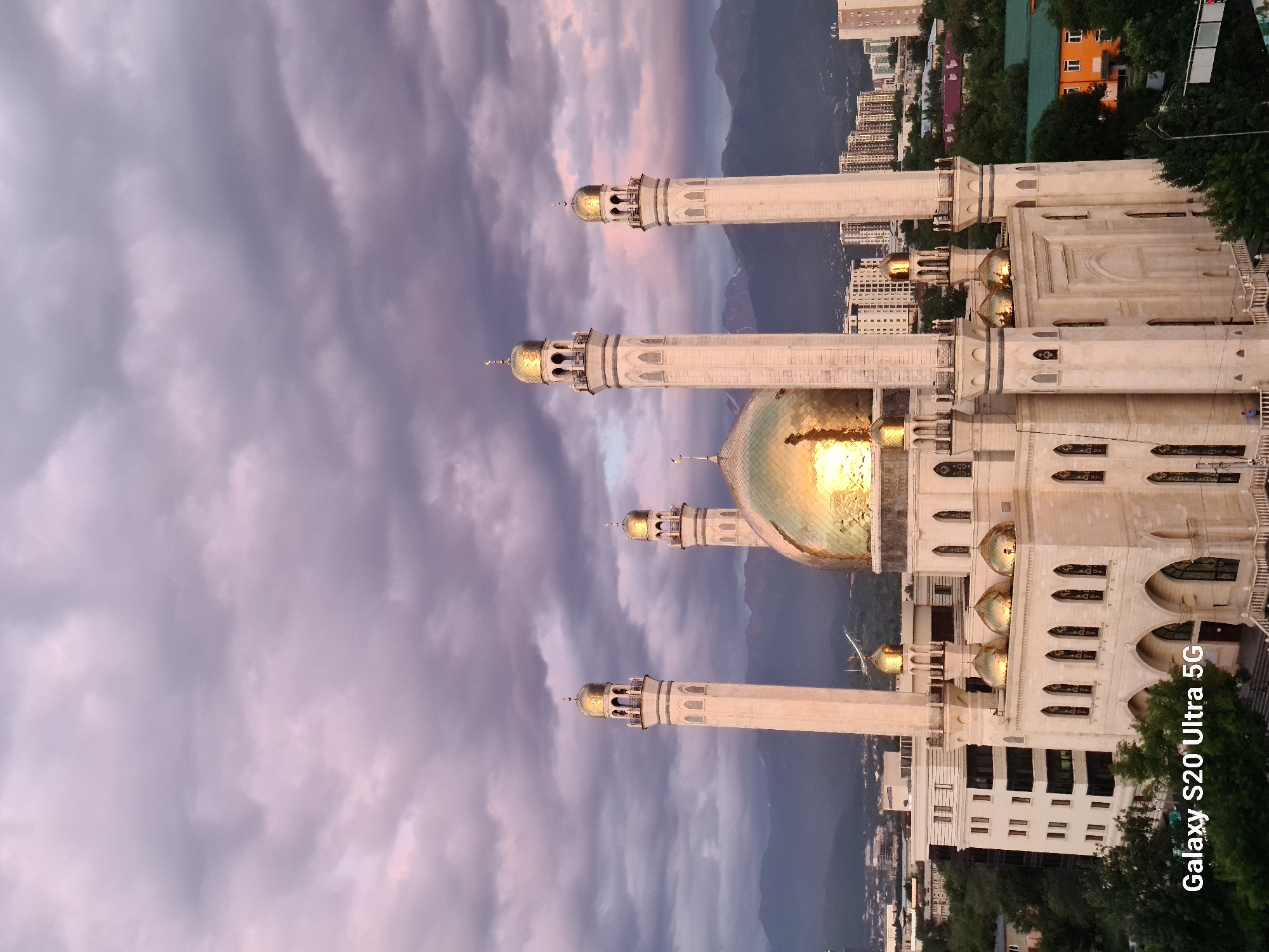 This is a vertically oriented image of the Central Mosque in Almaty, Kazakhstan, captured at dusk. The sky is dramatic with a mix of dark, purplish-grey clouds and streaks of pinkish-orange light, suggesting the sun has recently set or is about to rise, illuminating the clouds from below.
The main subject is the mosque, a grand structure with pale stone facades and multiple golden domes. Several tall, slender minarets with rounded tops and decorative balconies rise from the building. The largest, central dome is particularly striking, reflecting the vibrant colors of the sky. The mosque's architecture features arched windows and intricate decorative elements.
In the background, the city of Almaty unfolds, with numerous tall apartment buildings and other structures visible. Beyond the city, a range of mountains can be seen, shrouded in the twilight haze. Lush green trees are interspersed with the urban landscape and frame the lower part of the mosque.
The lighting and colors create a serene and majestic atmosphere. The time of day is clearly twilight, with the soft, diffused light casting a warm glow on the golden domes and highlighting the textures of the clouds.
Visible text in the lower right corner reads: "Galaxy S20 Ultra 5G".