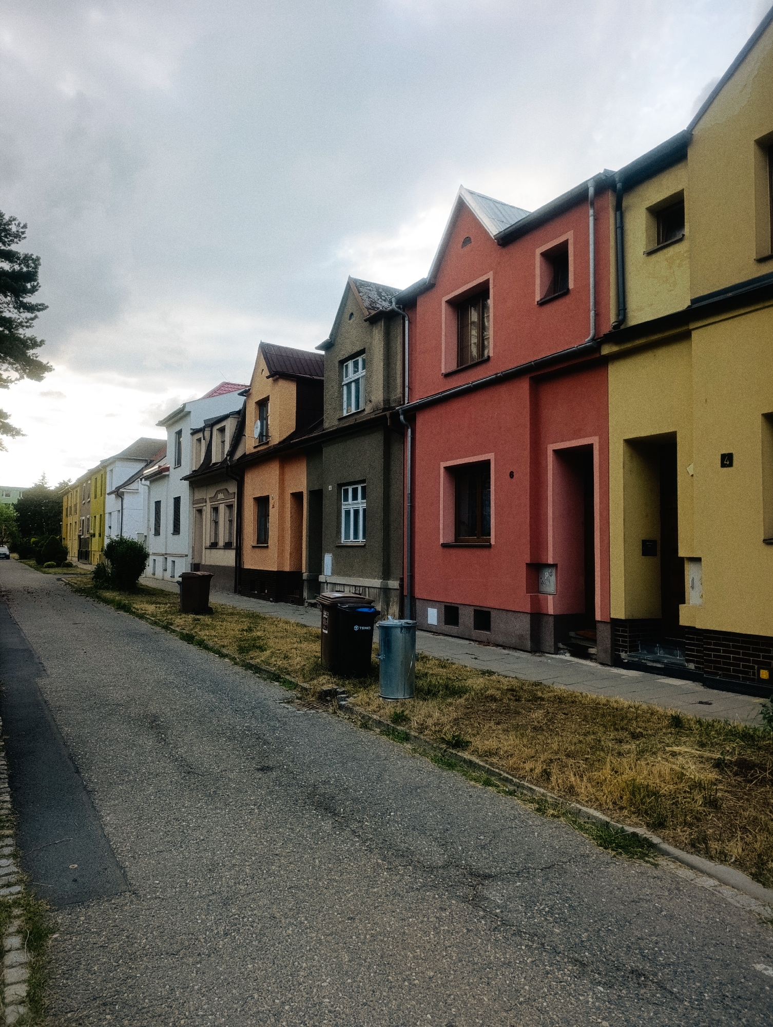 The image shows a row of colorful, two-story residential buildings lining a paved street in Olomouc, Czechia. The buildings are painted in varying shades of yellow, orange, and red. There are sidewalks in front of the buildings, and some dry grass and weeds grow alongside them. Several trash cans are visible on the sidewalk. The sky is overcast. The perspective is from the street, looking down the row of houses.