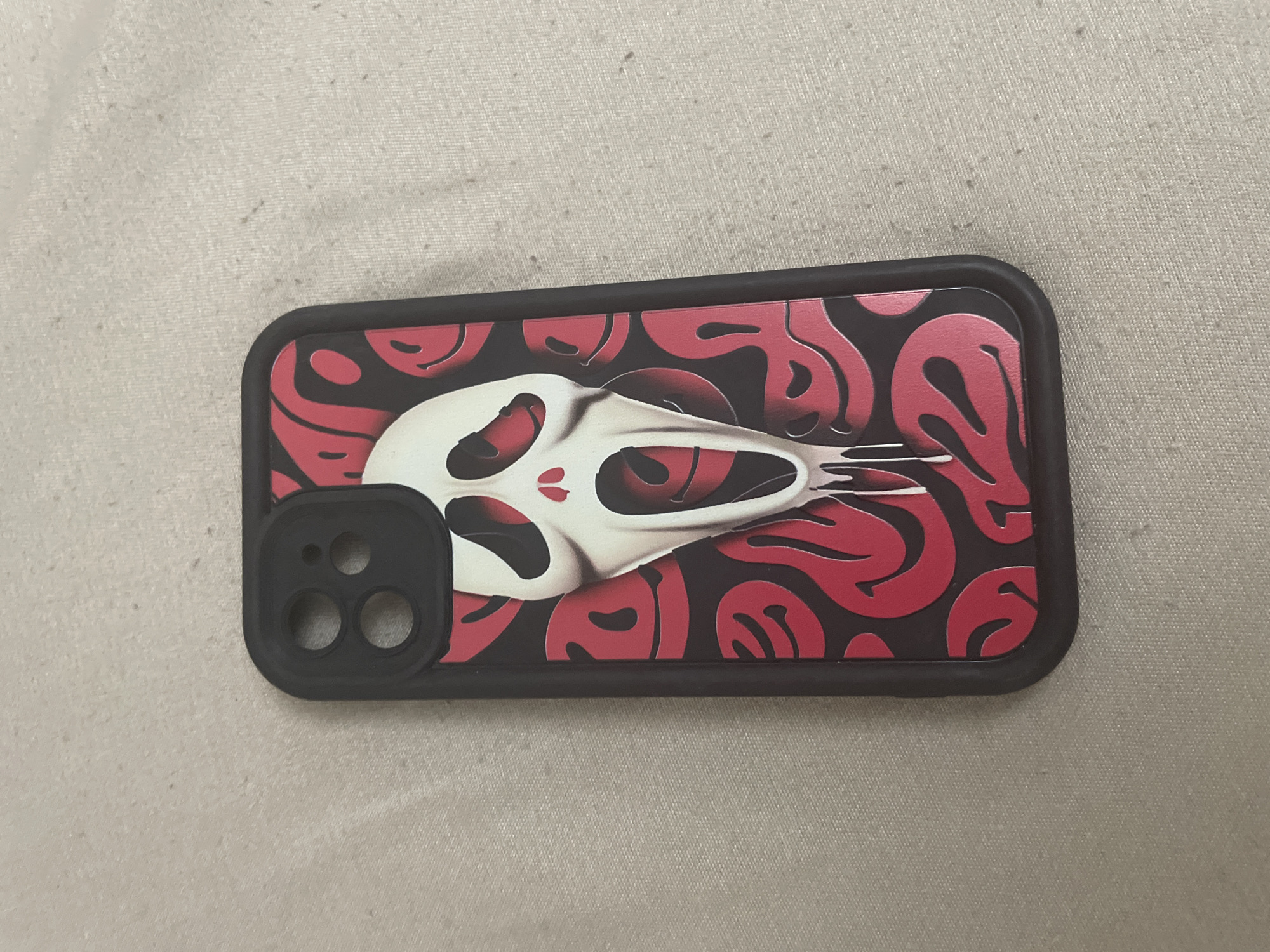 A black and red iPhone case is depicted lying face-up on a light-colored, subtly textured fabric surface. The case features a prominent design: a stylized white "Scream" ghost face mask positioned centrally. The mask has black, elongated eye and mouth openings, with two small red drips visible beneath the eye holes. The lower portion of the mask appears to be depicted as melting or dripping, forming irregular white streaks. The background of the case design consists of multiple distorted, wavy, red smiley face patterns on a dark grey or black base. The black frame of the phone case is visible around the perimeter of the design. The upper left corner of the case shows a black camera cutout section, designed for two camera lenses and an additional circular opening, likely for a flash or sensor. The fabric surface beneath the case exhibits a fine weave texture with various small, dark specks irregularly distributed across it. The exact location, including the city of Ihtiman, Bulgaria, cannot be determined from the provided image.