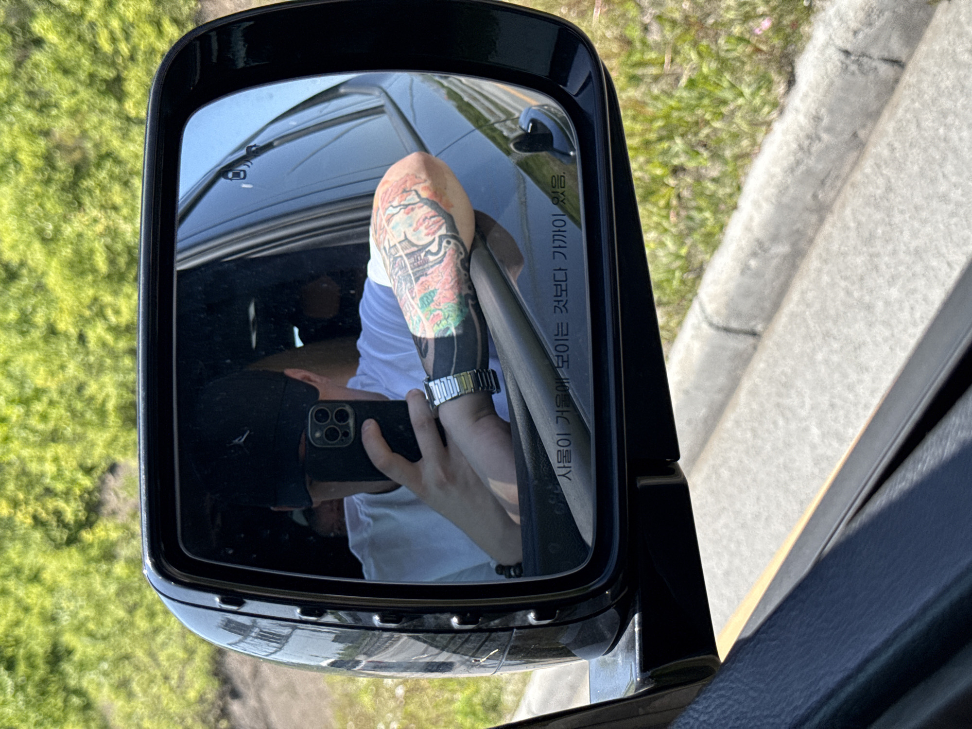 The image shows the side mirror of a dark-colored car reflecting a person holding a smartphone. The person is wearing a white shirt, a cap, and has a tattoo on their arm. The mirror is capturing the reflection of the person taking a photo or video. The background visible in the mirror includes the interior of the car, and outside the car, a green grassy area and a concrete wall or curb are visible. There is text in Korean on the mirror. The location is not specified and there is no information to suggest that the location is Vladivostok, Russia.