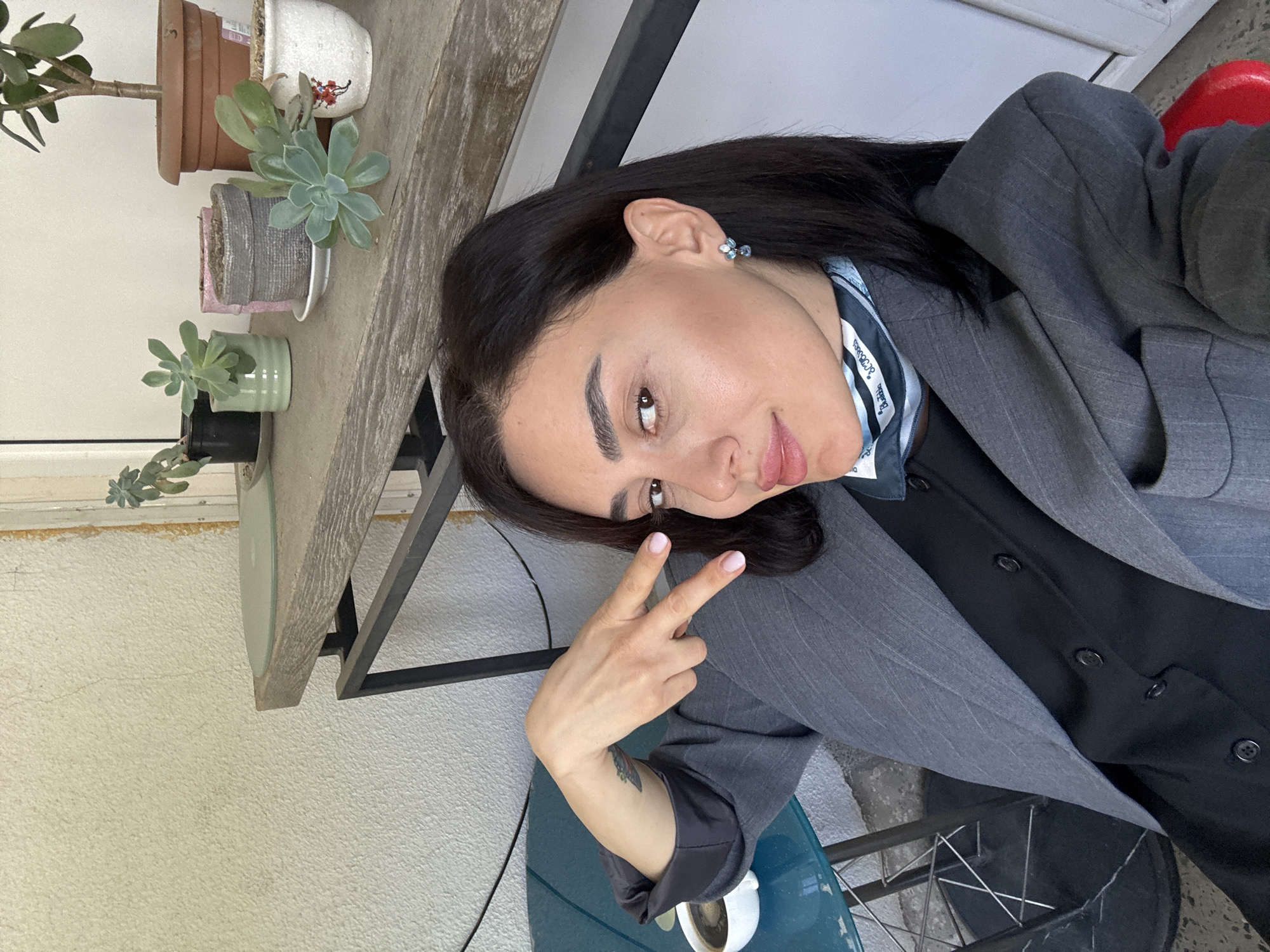The image is a selfie taken by a woman. She has dark hair, fair skin, and is wearing a gray blazer over a black shirt with a blue patterned scarf. She is holding up a peace sign with her right hand. A small tattoo is visible on her inner forearm.
Behind her, there is a weathered wooden shelf holding several potted plants, including succulents. The wall behind the shelf is off-white. The woman is sitting on a blue chair with a cup of coffee on the table next to it. In the background, there is a partial view of a red object. Based on these details, the location appears to be an outdoor seating area in Tbilisi, Georgia.