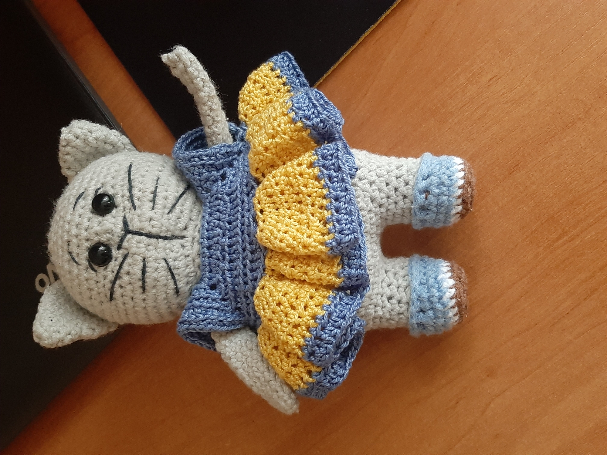 A crocheted stuffed cat toy stands upright on a wooden surface. The cat is light gray and wears a blue dress with a yellow ruffled skirt. It has black button eyes and embroidered whiskers and a nose. Its feet are covered in light blue crocheted booties with white trim. The background is out of focus, showing a dark surface with a diagonal line, possibly a desk or table. The scene is indoors.