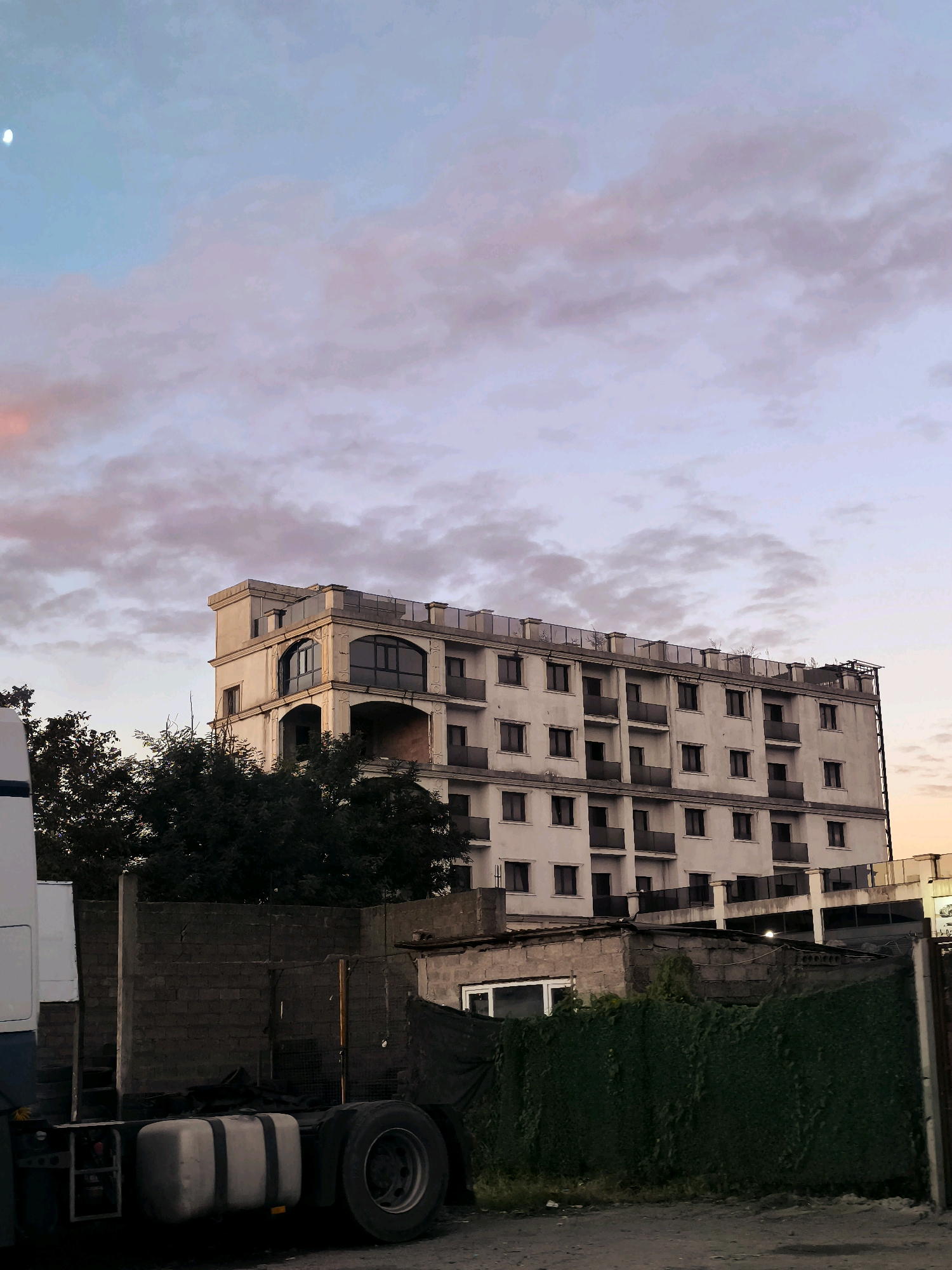 An exterior, eye-level, medium shot shows a building with multiple floors and a truck in the foreground, under a sky with clouds and a sliver of moon. The building is white and appears to be a hotel or apartment complex, with a modern architectural style and dark-tinted windows. The truck is partially visible on the left side of the frame, with its large wheels and fuel tank prominent. In the middle ground, there are some lower structures, possibly warehouses or garages, constructed from concrete blocks. A green fence covered in vines runs along the right side of the frame. The sky is a gradient of blue and pink, suggesting either sunrise or sunset. The overall scene depicts an urban environment, with a mix of modern and utilitarian structures.