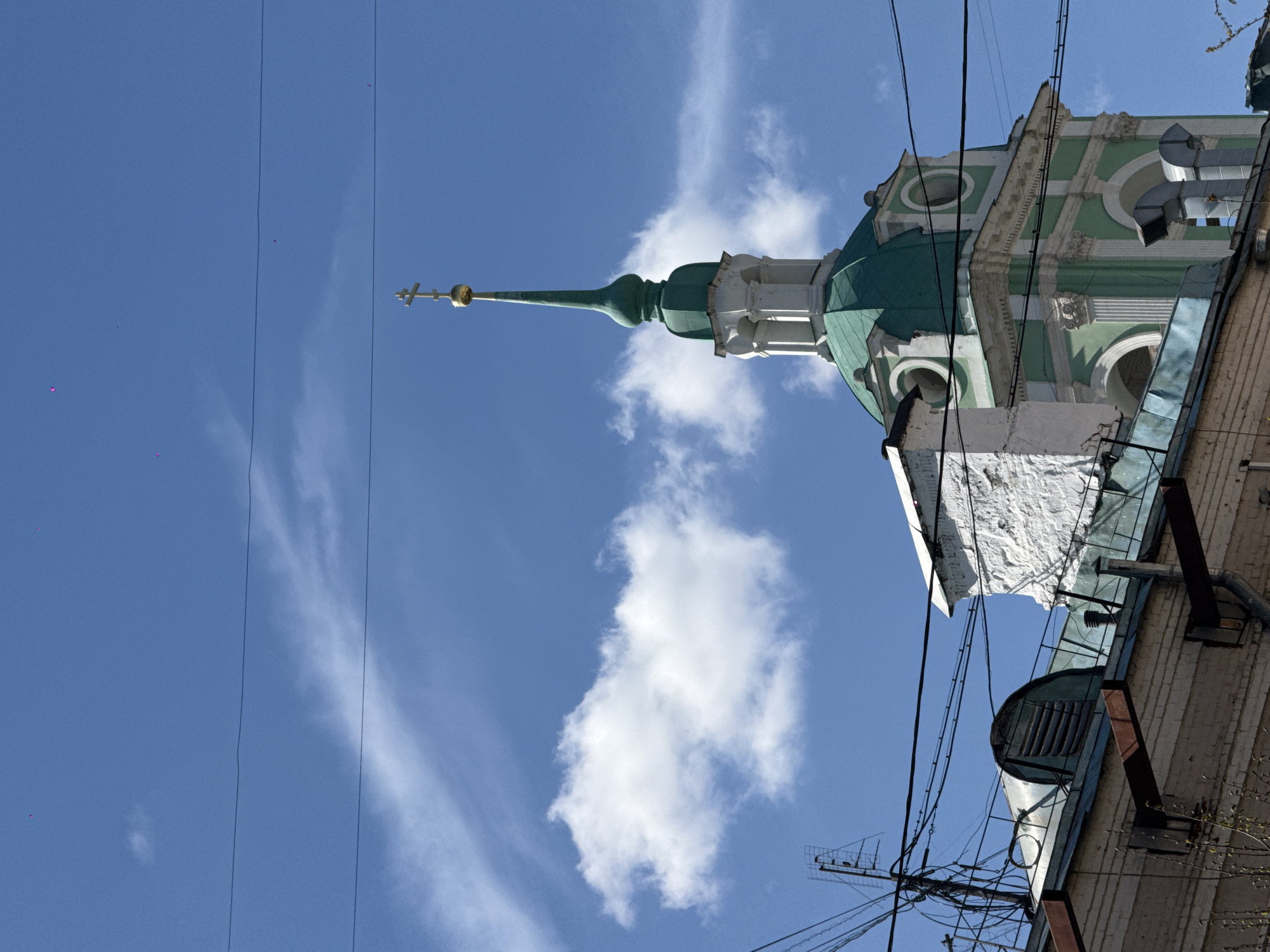 The image depicts a low-angle view of a building in Moscow, Russia, featuring a prominent Orthodox church spire against a blue sky with scattered clouds. The spire is painted in shades of green and white, topped with a golden cross. Parts of the building adjacent to the church spire are visible, including brickwork, metal fixtures, and a ventilation system. Overhead power lines run across the sky, adding to the urban setting.