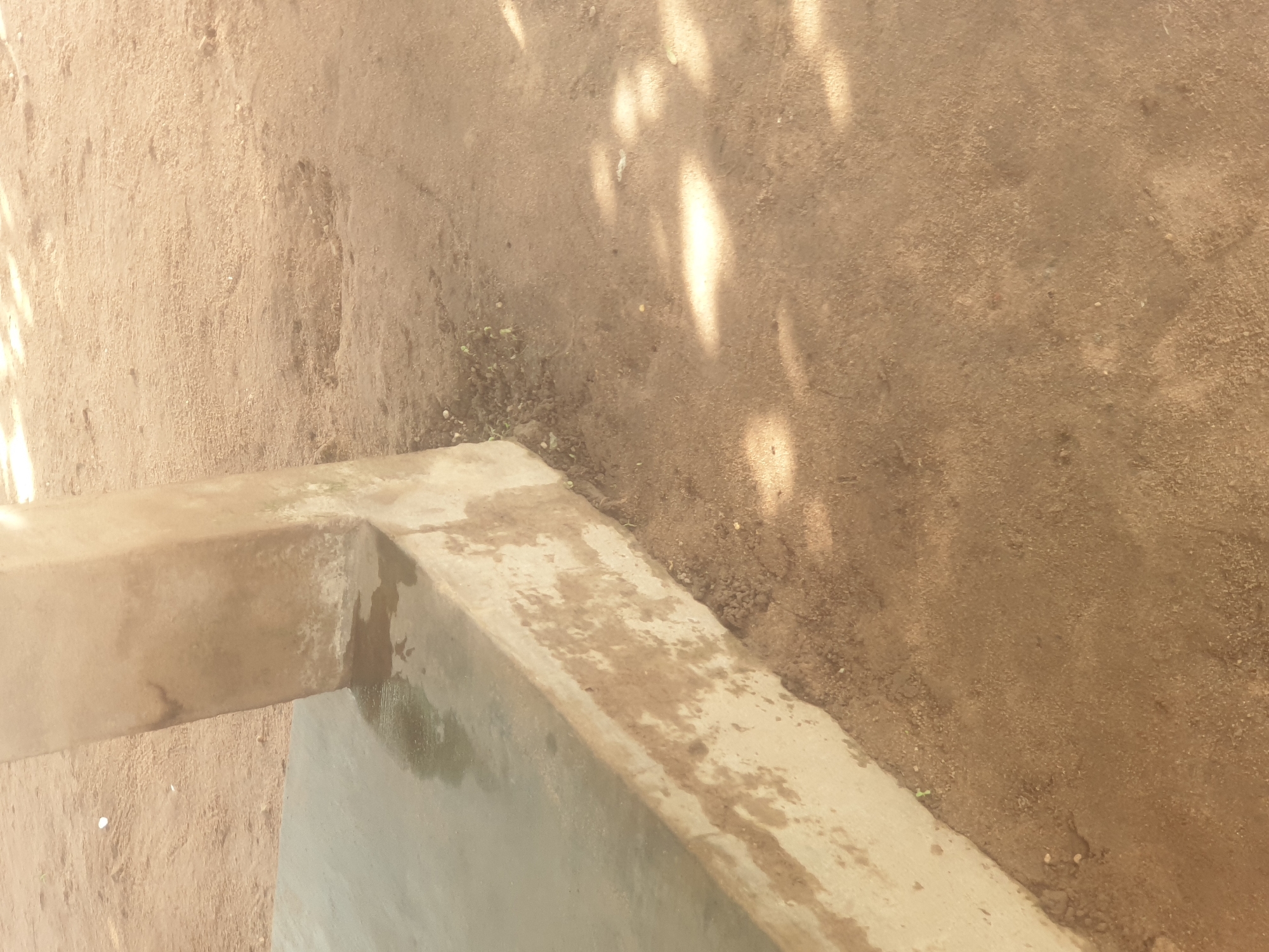 The image depicts a concrete structure and surrounding sandy ground, consistent with an outdoor area in Jauro Yinu, Nigeria. The concrete appears to be part of a raised platform or foundation, with a wet surface indicating recent water presence. The ground is dry and sandy, with some scattered debris and small patches of sparse vegetation. The lighting suggests it is daytime, with dappled shadows on the sand, likely from overhead foliage. There are no people or animals visible in the frame. The overall impression is one of an unadorned, utilitarian outdoor space.