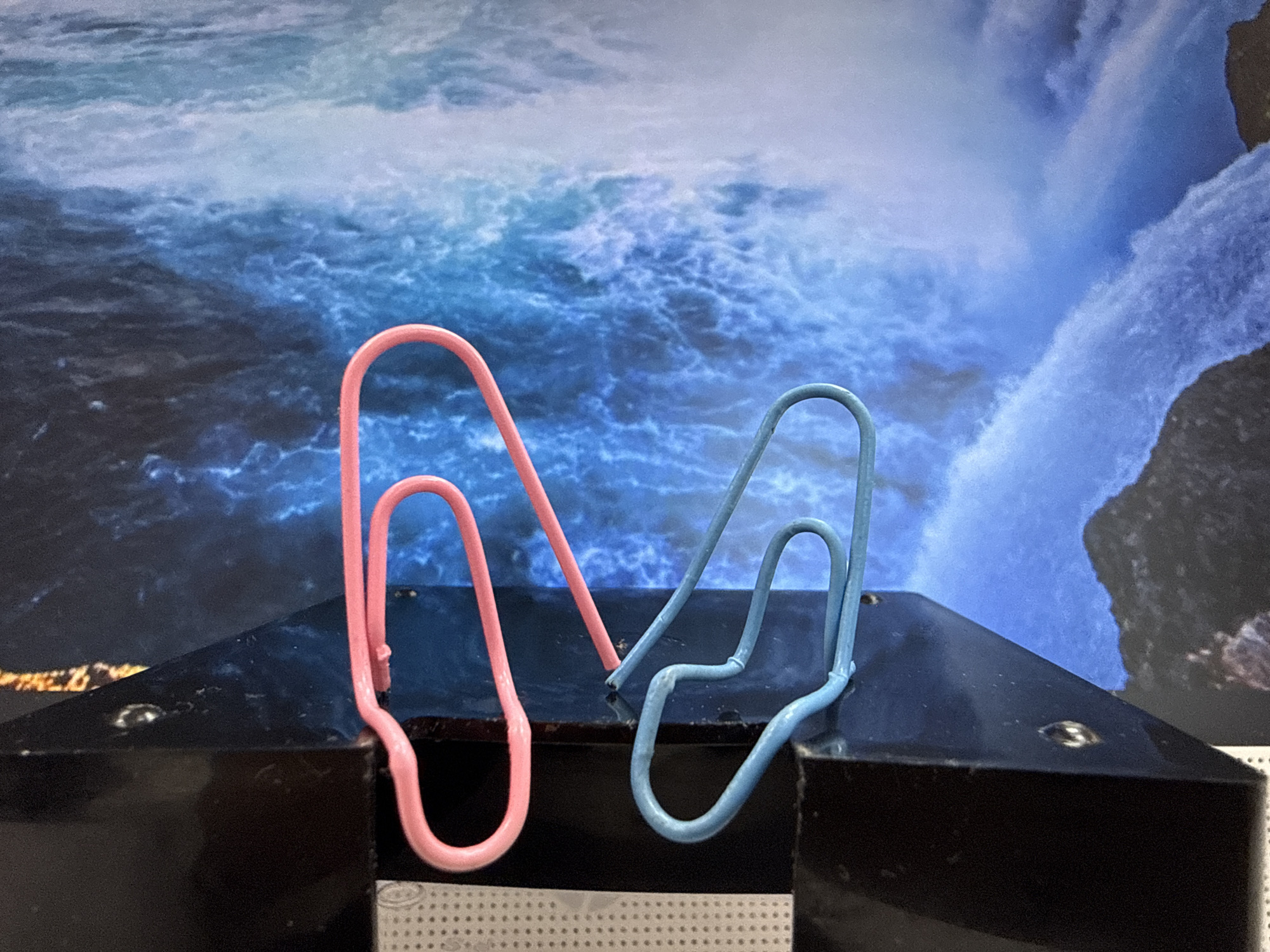 A close-up shot depicts two paperclips, one pink and one light blue, standing on a glossy black surface. The background features a printed image of turbulent, blue water and a cascading waterfall. The lower part of the glossy surface shows a partially visible white grid pattern. While the image provides no direct evidence of location, the question explicitly mentions the city of Chaikovsky, Russia. Therefore, it can be inferred that the scene potentially originates from or is representative of an environment within Chaikovsky, Russia, although confirmation is not possible based solely on visual data.