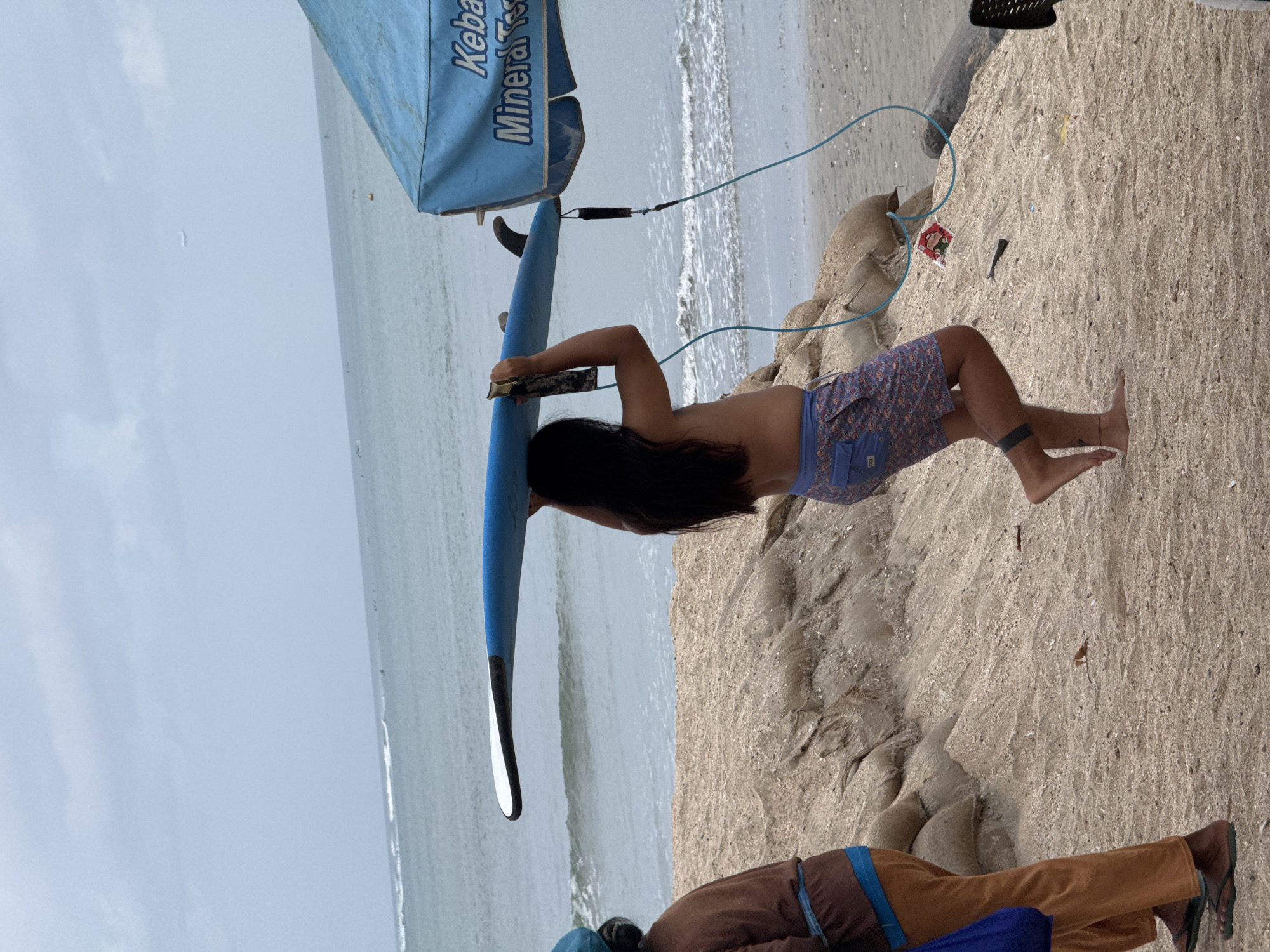 In Kuta, Indonesia, a person with long, dark hair walks on a sandy beach, carrying a blue surfboard horizontally above their head. They are wearing patterned shorts. To the left, another person walks away from the camera, wearing brown pants and a blue top underneath a brown outer layer, carrying a blue bag. In the background, the ocean is visible with waves and several surfers in the distance. A blue canopy with text that includes "Keba" and "Mineral" stands on the right side of the frame, partially obscuring the view of the ocean. The beach is composed of sand and small shells, with sandbags piled on a slope behind the people. The sky is overcast.