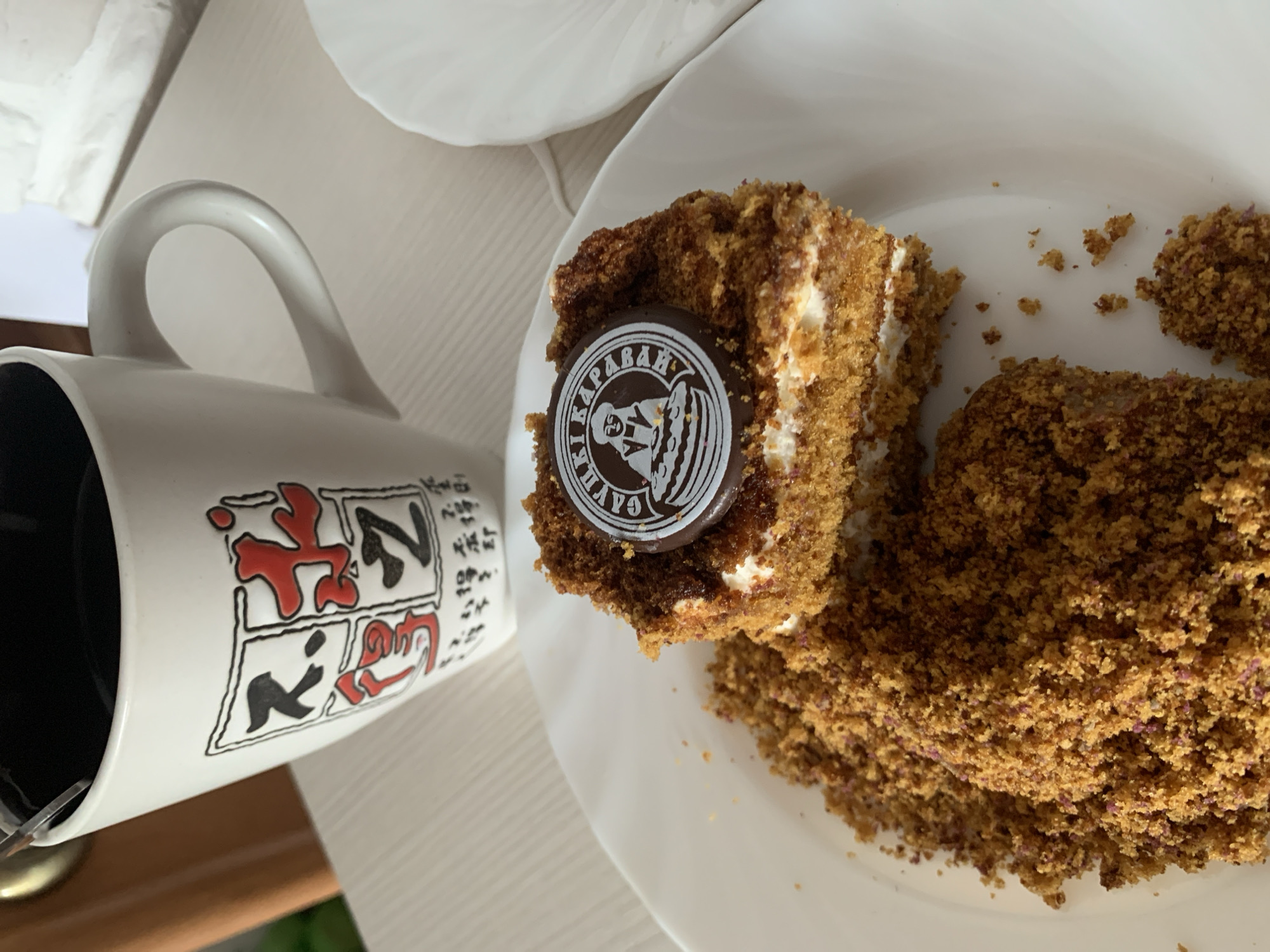 The image shows a slice of cake on a white plate next to a white mug. The cake is a layered pastry, possibly a honey cake, covered in crumbs. A small, round, dark chocolate disc with a white emblem is placed on top of the cake. The emblem features a silhouette of a person with a crown and intricate designs. The mug is white with black and red illustrations of what appear to be Japanese characters. The scene is set on a light-colored, textured surface, possibly a table, and part of another white plate is visible in the background.
The lighting suggests it is daytime. There are no specific weather cues. The presence of the cake and coffee in a mug implies a casual setting, possibly a cafe or a home breakfast/dessert setting. The overall mood is relaxed and inviting, suggesting enjoyment of a sweet treat and a beverage.
Visible text on the chocolate disc appears to read "КАРАВАЙ" (Karavai) in Cyrillic, which is also the name of a popular bakery chain in Belarus. The characters on the mug are in Japanese Kanji and Hiragana.