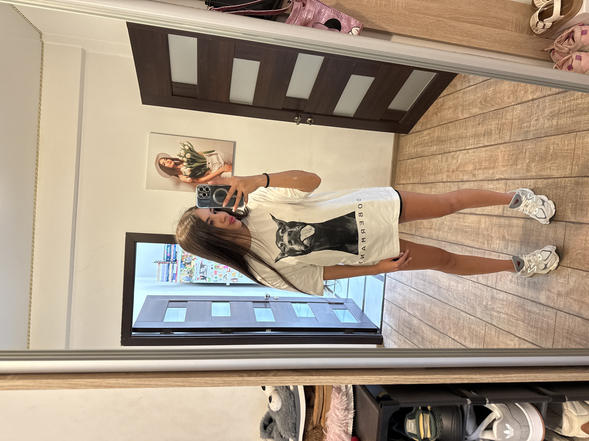 A young woman with long brown hair stands in front of a full-length mirror, taking a selfie with her phone. She is wearing an oversized white t-shirt with a black graphic of a doberman dog sticking its tongue out and black shorts. She has on white and grey athletic shoes. Her reflection shows her in what appears to be an indoor hallway with wood-paneled walls and floor. To her left, a shoe rack is visible, stacked with various shoes. To her right, a dark brown door with glass inserts is prominent. In the background, reflected in the mirror, is a framed picture of a woman holding flowers and another picture with various colorful images. The overall environment suggests a residential interior.