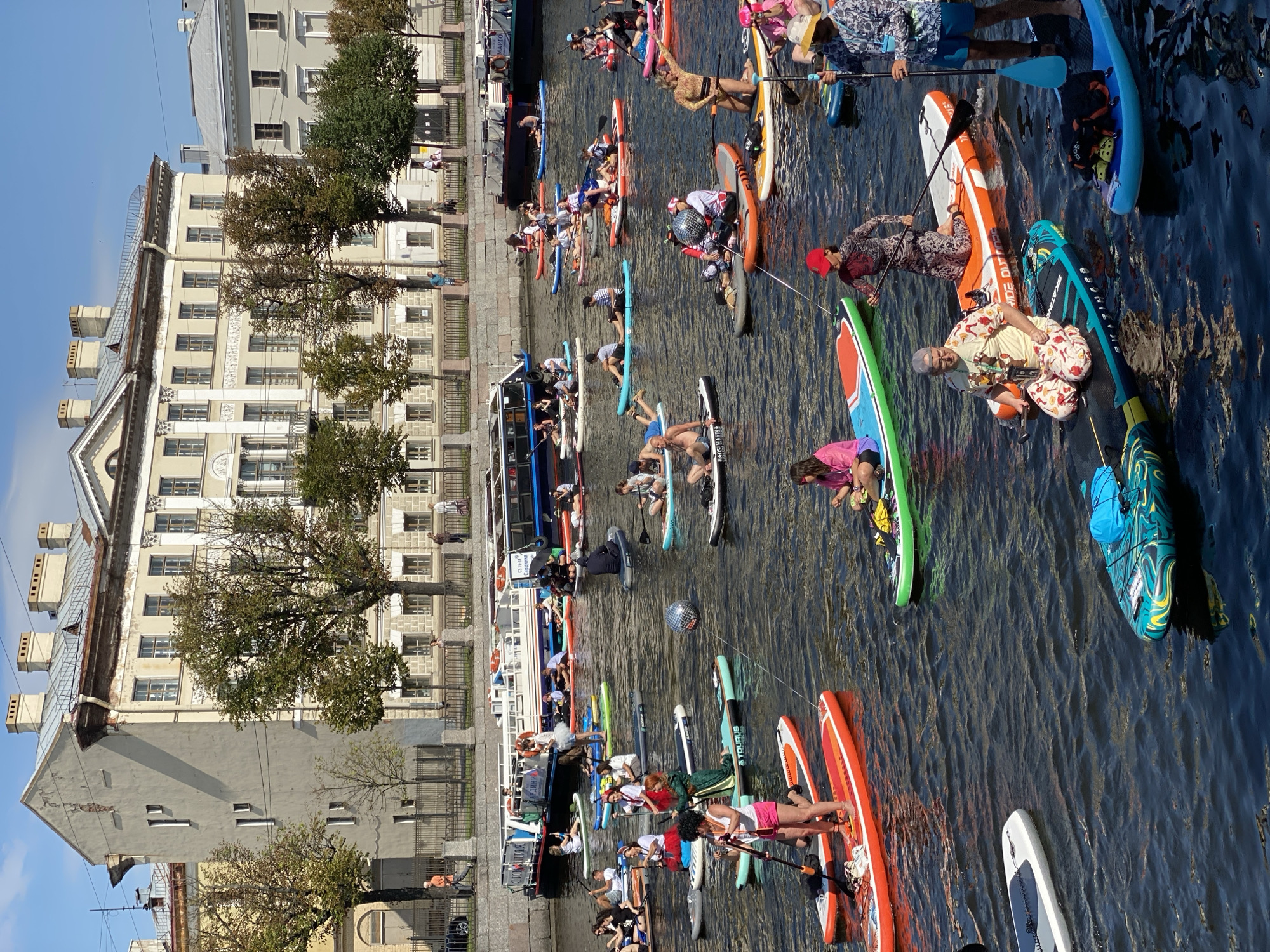 In Saint Petersburg, Russia, a group of people are paddleboarding on a canal. Several buildings line the canal, and trees are visible along the banks. The paddleboarders are diverse in age and gender, and many are dressed in colorful outfits, some in costumes. Some are paddling, while others are sitting or kneeling on their boards. A disco ball is suspended in the air, and a few boats are docked in the background. The water is dark and choppy, reflecting the blue sky and the surrounding architecture.