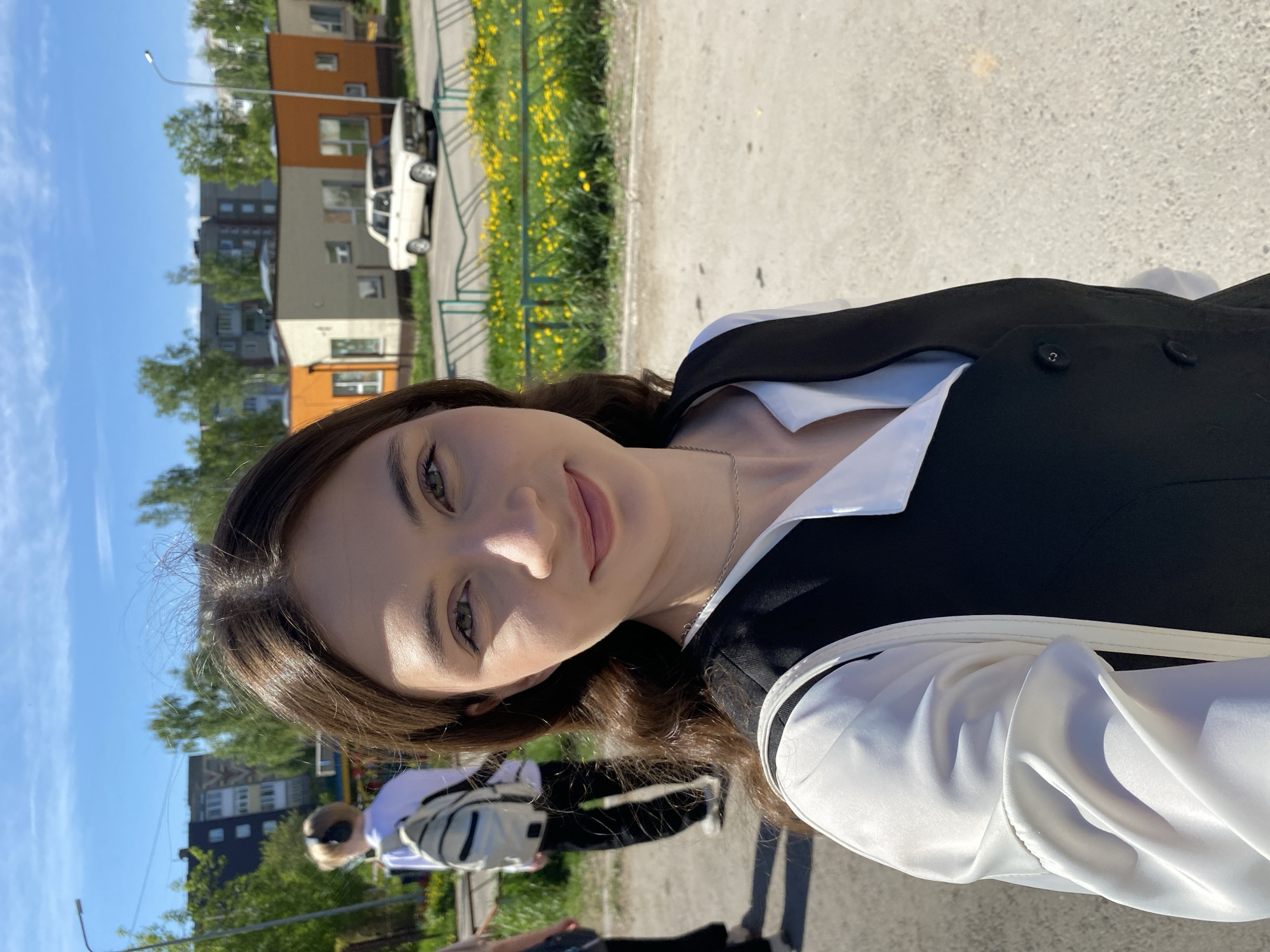 A young woman is shown from the waist up in a selfie shot outdoors on a sunny day. She has long, dark hair and is wearing a white blouse with a black vest. The background includes a city street with apartment buildings and yellow flowers near a fence. A white car is visible in the background, and another person is partially visible further behind. The general location is Votkinsk, Russia, based on the apartment architecture, car model, and landscape.