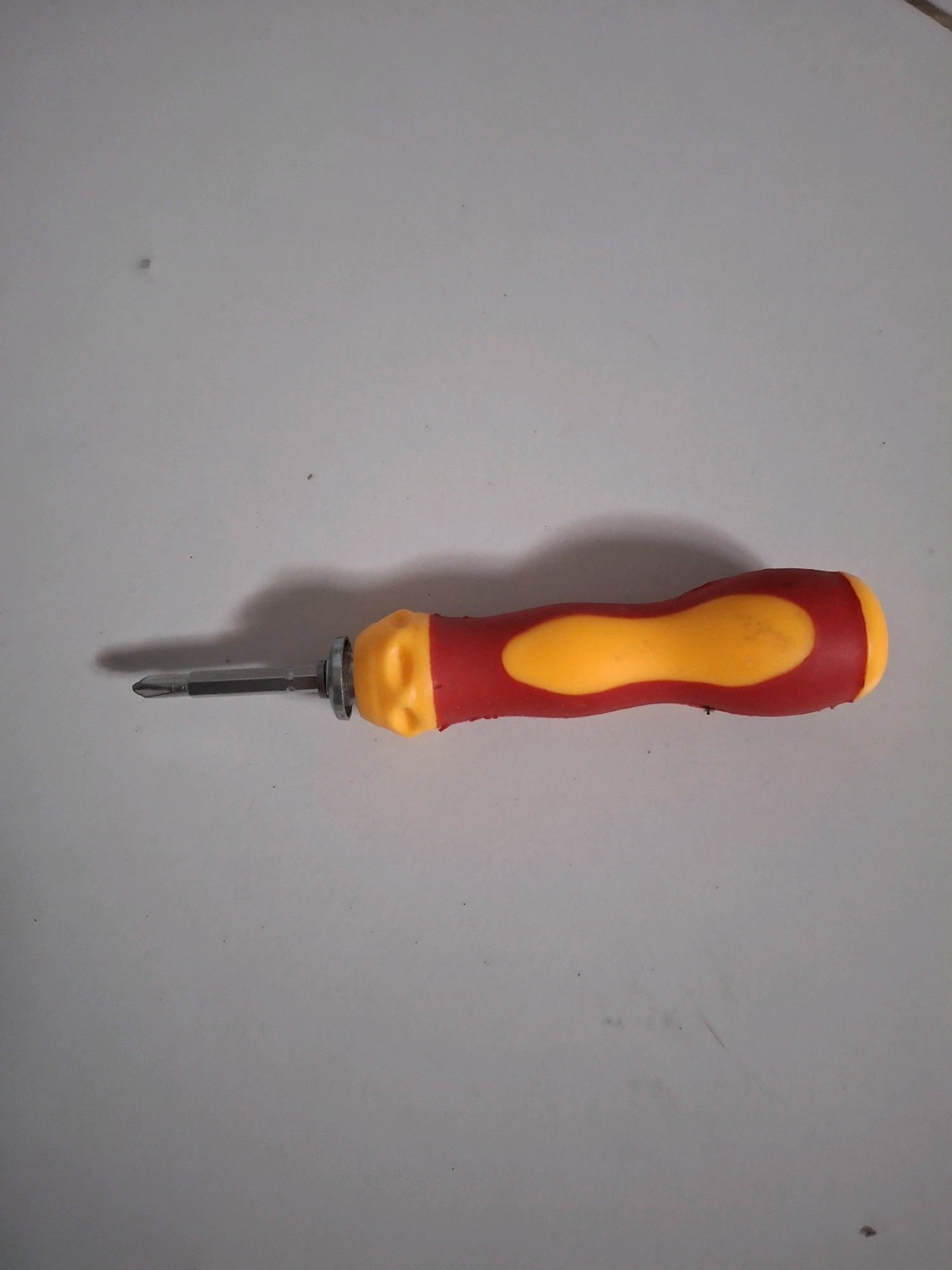 A Phillips head screwdriver with a red and yellow handle is placed horizontally on a white surface. The screwdriver is oriented from left to right, with the metal shaft and tip on the left and the handle on the right. The surface appears to be a flat, light-colored material, possibly a tabletop or floor. The lighting casts a slight shadow to the left of the screwdriver. The image was taken in Lumajang, Indonesia.