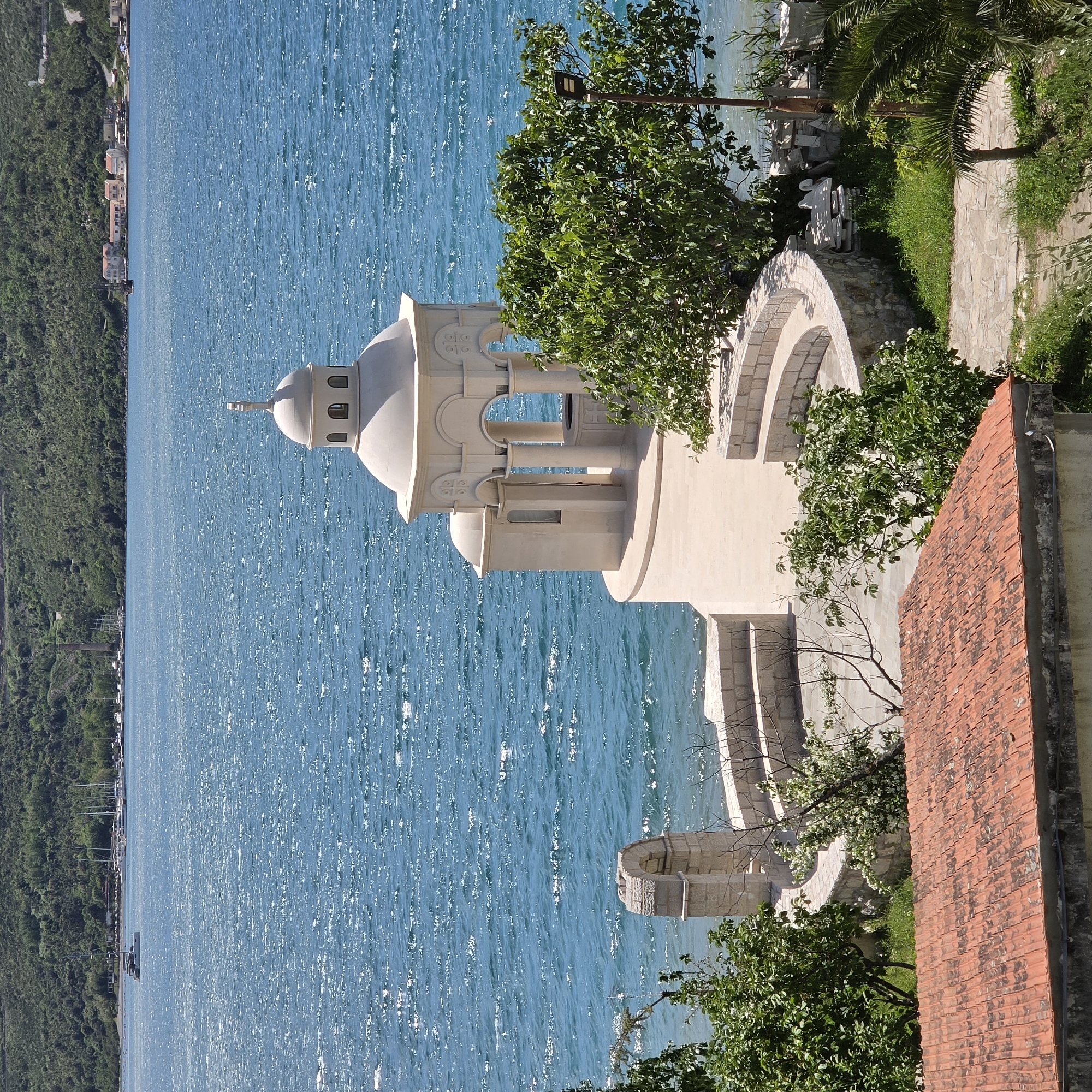 The image showcases a serene coastal scene in Đuraševići, Montenegro. A white domed building, possibly a church or religious structure, stands prominently in the foreground against the backdrop of a sparkling blue sea. The architectural style includes arches and decorative columns. Lush green trees and other vegetation surround the structure, softening the lines of the building. A red-tiled roof suggests a neighboring building or structure. The sea stretches out towards the horizon, with the distant coastline marked by green mountains. The sky is clear, suggesting a sunny day.