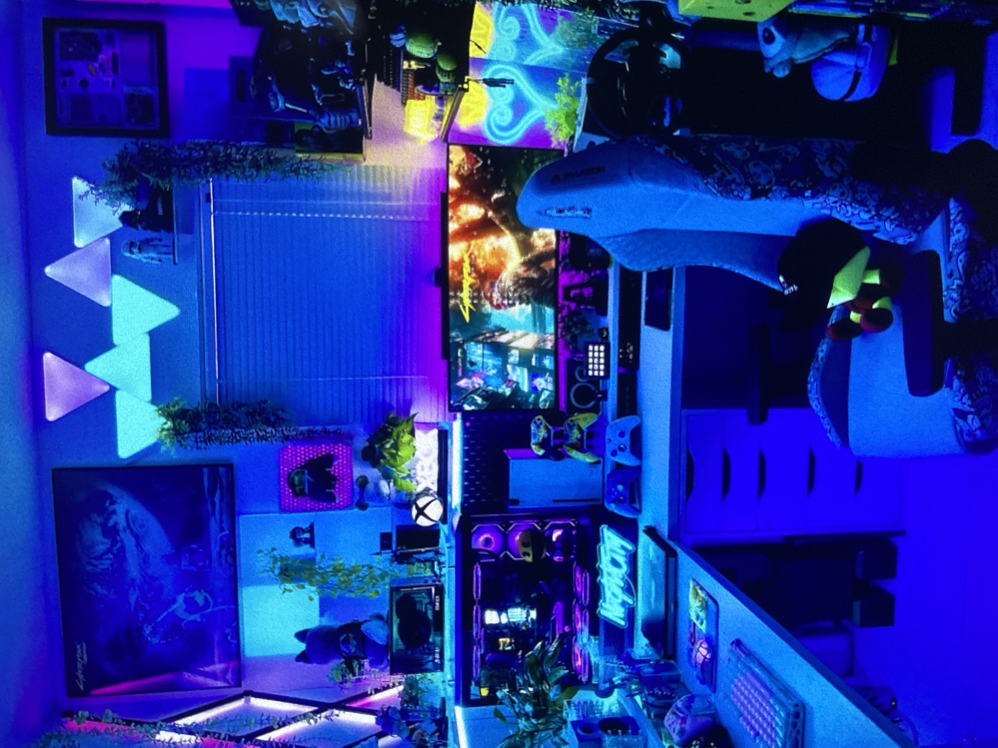 The image shows a gaming setup in a room illuminated primarily by blue and purple lighting. The setup includes multiple monitors displaying various images and game scenes. A gaming chair is visible to the right, with what appears to be a plush toy resting on it. In the background, there are shelves or display areas filled with various objects, including plants, posters, and gaming accessories. A "Night City" neon sign is also visible near the gaming computer. No people are visible. There's no indication of the location being Lhokseumawe, Indonesia.
