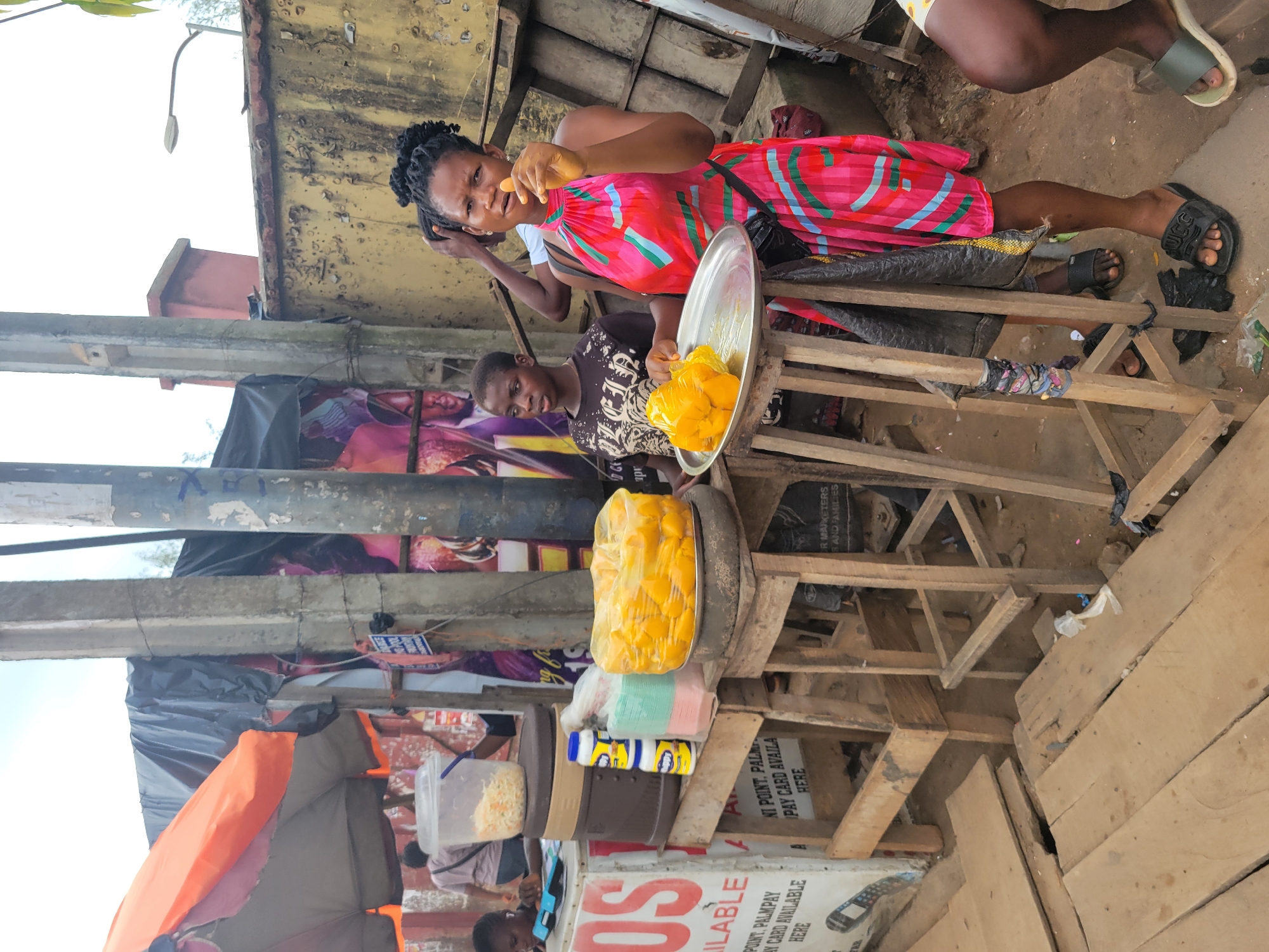 In Egbagu, Nigeria, a young woman in a pink and green patterned dress stands at a market stall, holding a tray of what appears to be mangoes. She is looking towards the camera and has her right hand to her mouth, possibly tasting the fruit. Beside her, a young boy in a black t-shirt looks to the left. The stall features a variety of items, including a plastic container with what looks like shredded cabbage, bottles of what appears to be a liquid condiment, and stacks of colorful plastic containers. In the background, a black tarp is draped over structures, and posters are visible on a wall. The scene is outdoors, with a rough, wooden surface in the foreground.