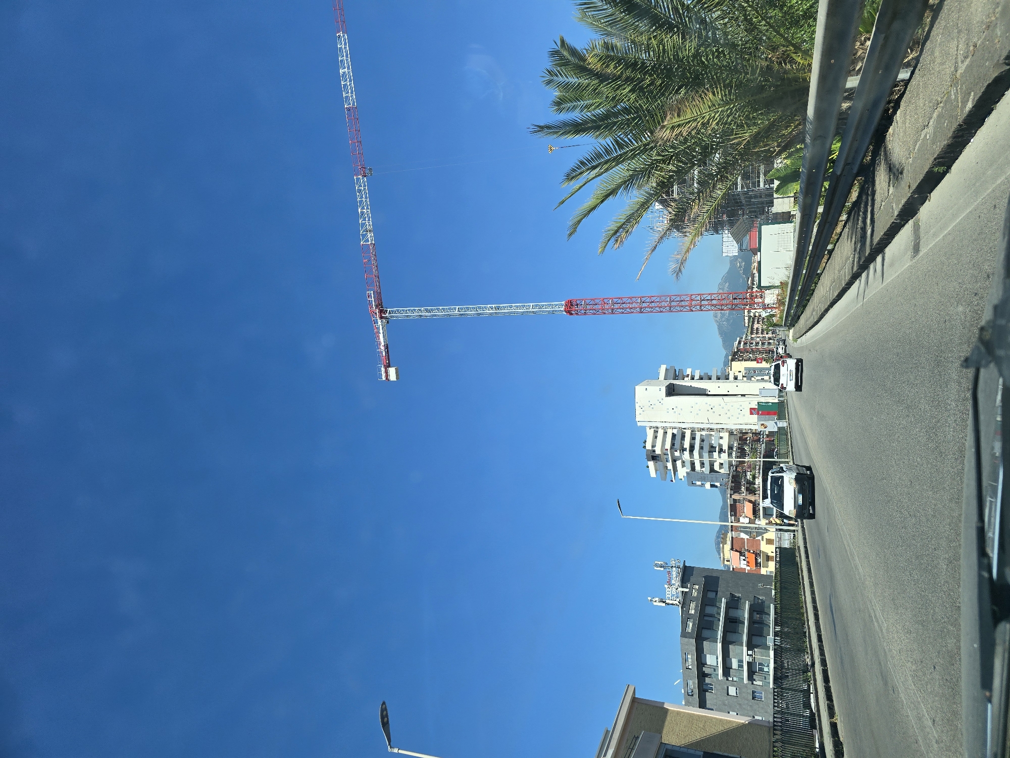 The image captures a view from a vehicle driving on a road in Salerno, Italy. A tall red and white construction crane dominates the sky on the right side of the frame. In the middle ground, several modern apartment buildings are visible, some with balconies and distinctive architectural features. A palm tree is prominently situated in the foreground on the right, partially obscuring the construction site. The road itself is asphalt, with a concrete barrier and sidewalk on the right. Two white cars are driving on the road, with the leading car being a small hatchback. The sky is a clear, bright blue, suggesting a sunny day. The overall scene depicts a developing urban area with ongoing construction.
