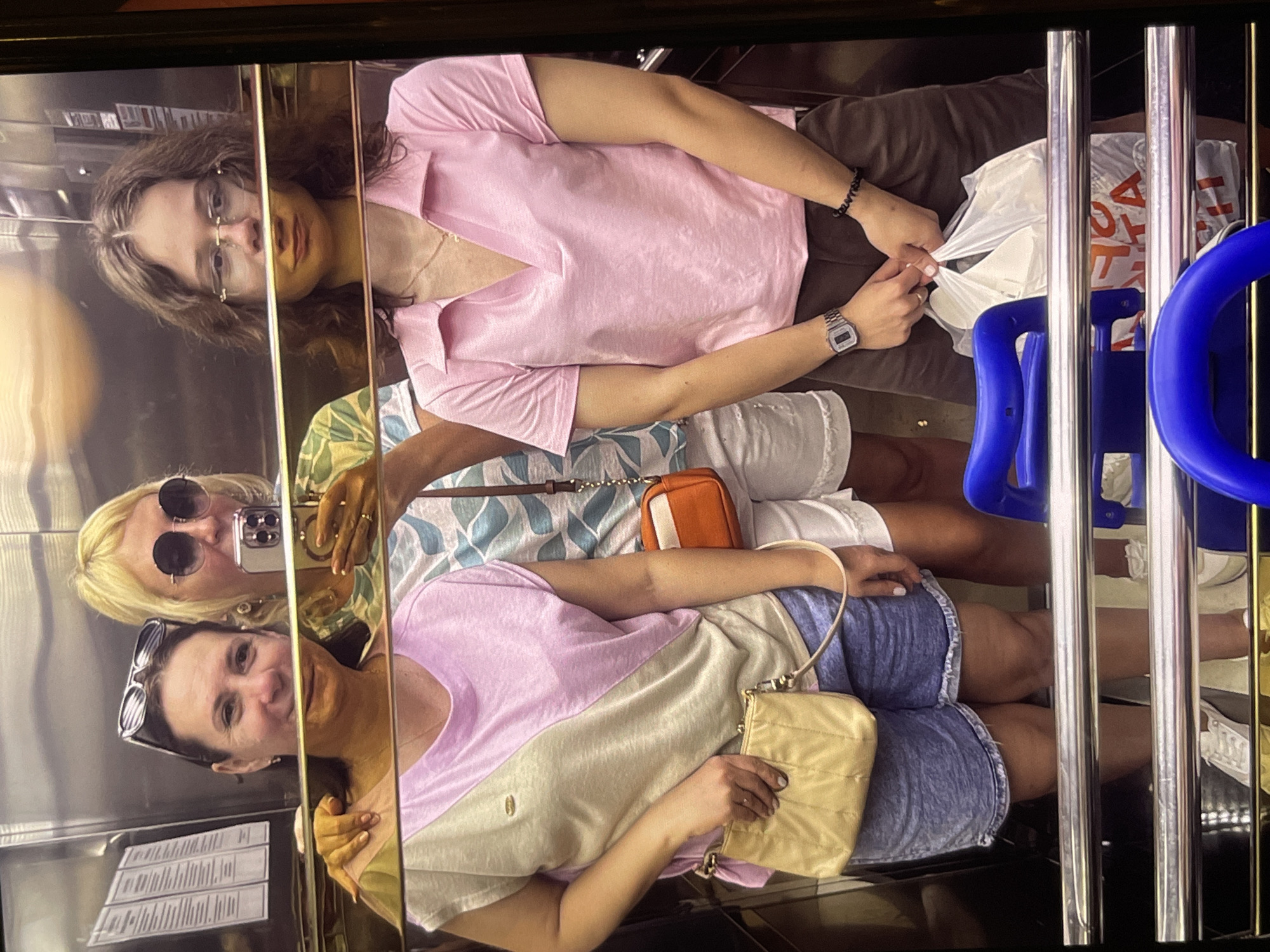 The image shows a selfie taken inside an elevator. Three women are reflected in the elevator's mirrored walls. One woman is positioned in the foreground wearing sunglasses on her head and a pink and beige shirt with denim shorts. Another woman, wearing round sunglasses and a patterned shirt, is holding a smartphone, presumably taking the photograph. A third woman is standing behind them, wearing glasses and a pink shirt. A blue child seat, and a plastic bag with red letters are visible within the elevator. The location is Unknown, Unknown.