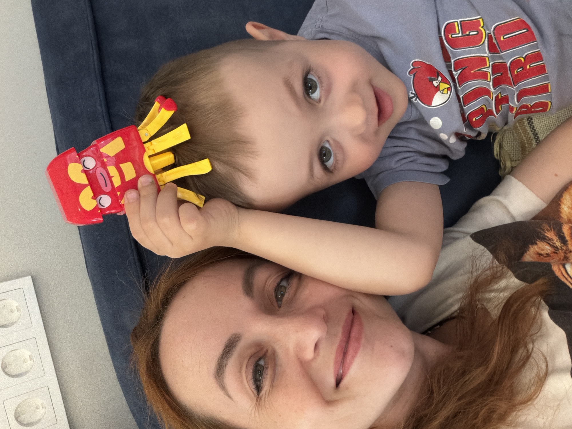 A woman and a young child are lying down. The child is wearing a light purple shirt with an "Angry Birds" graphic. The child is holding a red and yellow toy with protrusions. The woman is smiling. The background suggests they are on a blue upholstered surface, possibly a sofa. The image was taken indoors, in Chaikovsky, Russia.