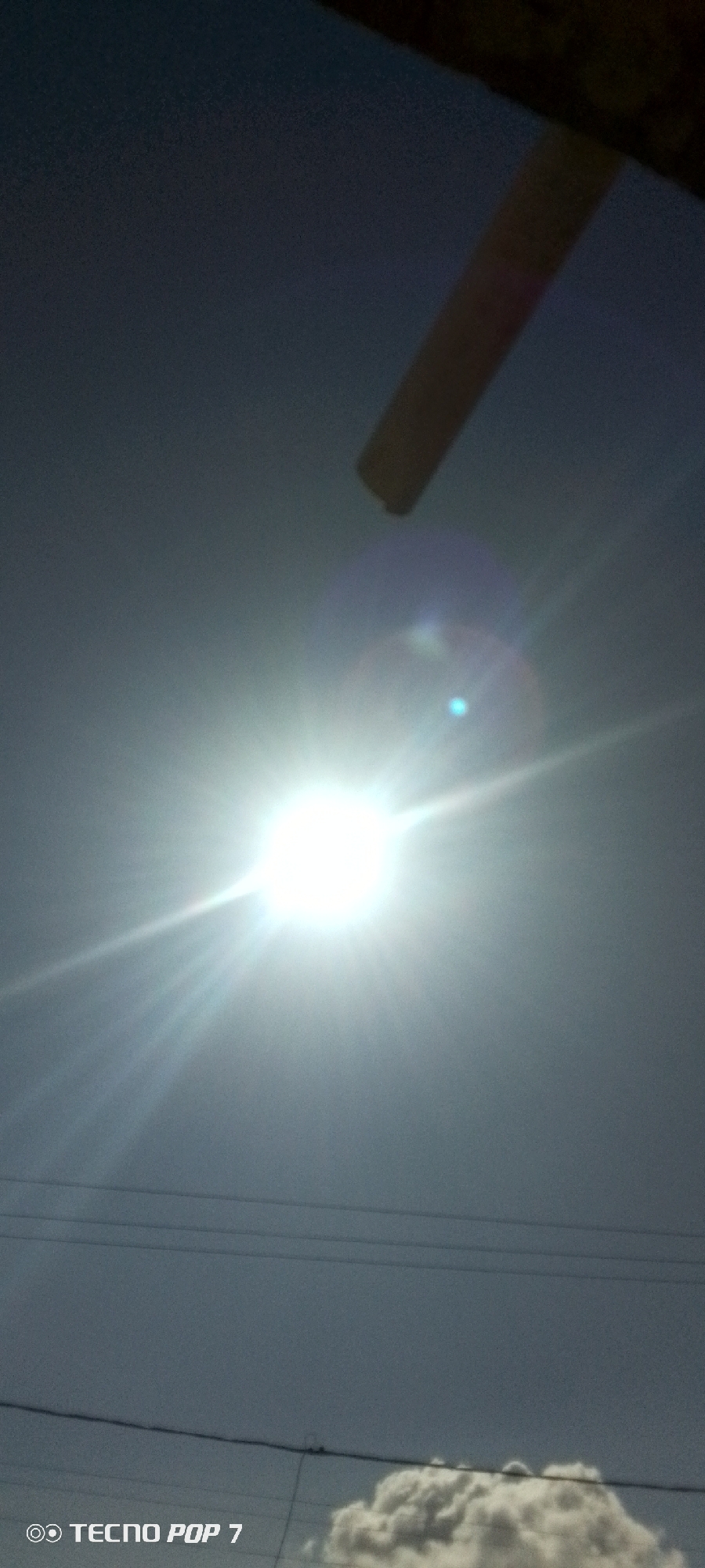 The image depicts a bright sun in a light blue sky, possibly Altagracia, Venezuela. Power lines are visible in the lower portion of the frame. A small cloud is present in the bottom right corner. A portion of a structure is visible in the upper right. The image was taken with a TECNO POP 7.