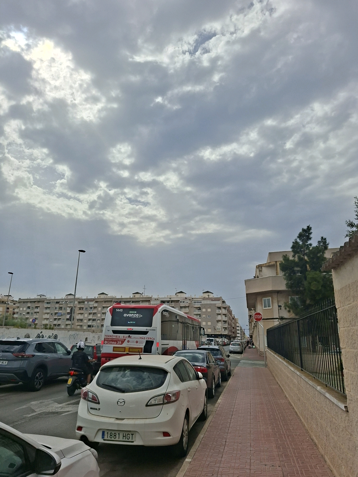 A street scene in Torrevieja, Spain, features a cloudy sky overhead. In the foreground, a white Mazda sedan with license plate "1881 HGT" is parked on the right side of the street, facing away from the camera. To its left, a dark SUV and a scooter with a rider wearing a white helmet are also visible. A red and white Avanza bus, numbered 1445, is stopped in traffic. Across the street, a long, multi-story residential building with numerous windows lines the background. To the right of the street, a paved sidewalk with a red brick pattern runs alongside a brick wall topped with a black metal fence. A red stop sign is visible on a building facade further down the sidewalk.