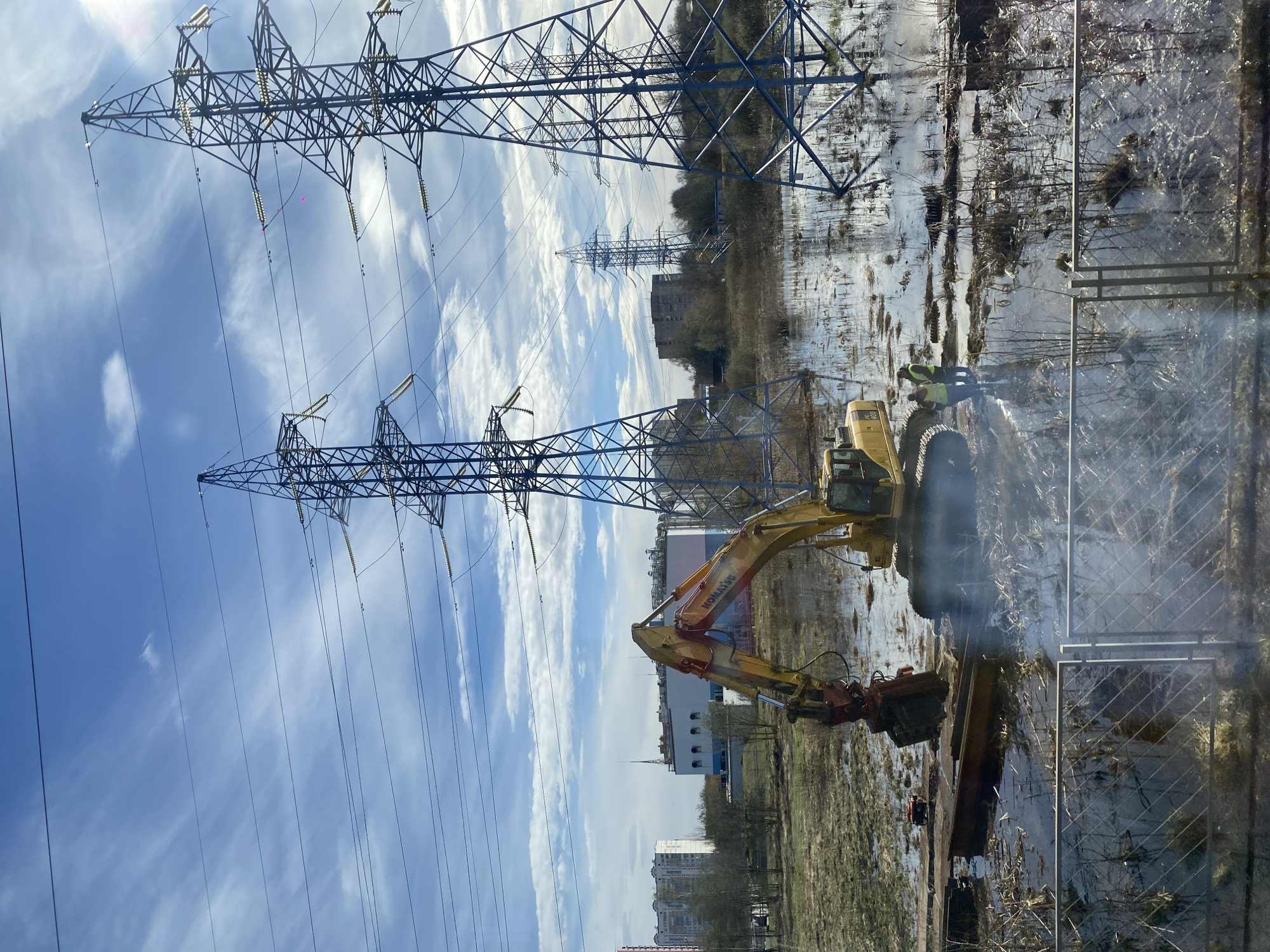 The image displays an active construction or industrial scene in an outdoor setting, likely in Moscow, Russia, under a partly cloudy sky during daylight hours. The dominant features are several towering metal electricity pylons, extending across the landscape and supporting numerous power lines.
In the foreground and midground, a large yellow Komatsu excavator is prominently positioned in a heavily flooded and muddy area. The excavator is equipped with a hydraulic breaker or similar attachment, suggesting demolition, breaking, or extensive digging work. One individual wearing a high-visibility yellow vest is visible near the excavator, possibly supervising or operating the machinery. Another less distinct figure is also present. The ground is characterized by standing water, mud, and sparse, bare vegetation, indicating a wet or marshy environment.
Beyond the immediate work area, there are sections of fencing and patches of bare trees. In the distance, various buildings are visible, including residential apartment blocks and what appear to be industrial or commercial structures, situating the scene within a semi-urban or industrial periphery. The brand name "KOMATSU" is clearly visible on the side of the excavator's arm.