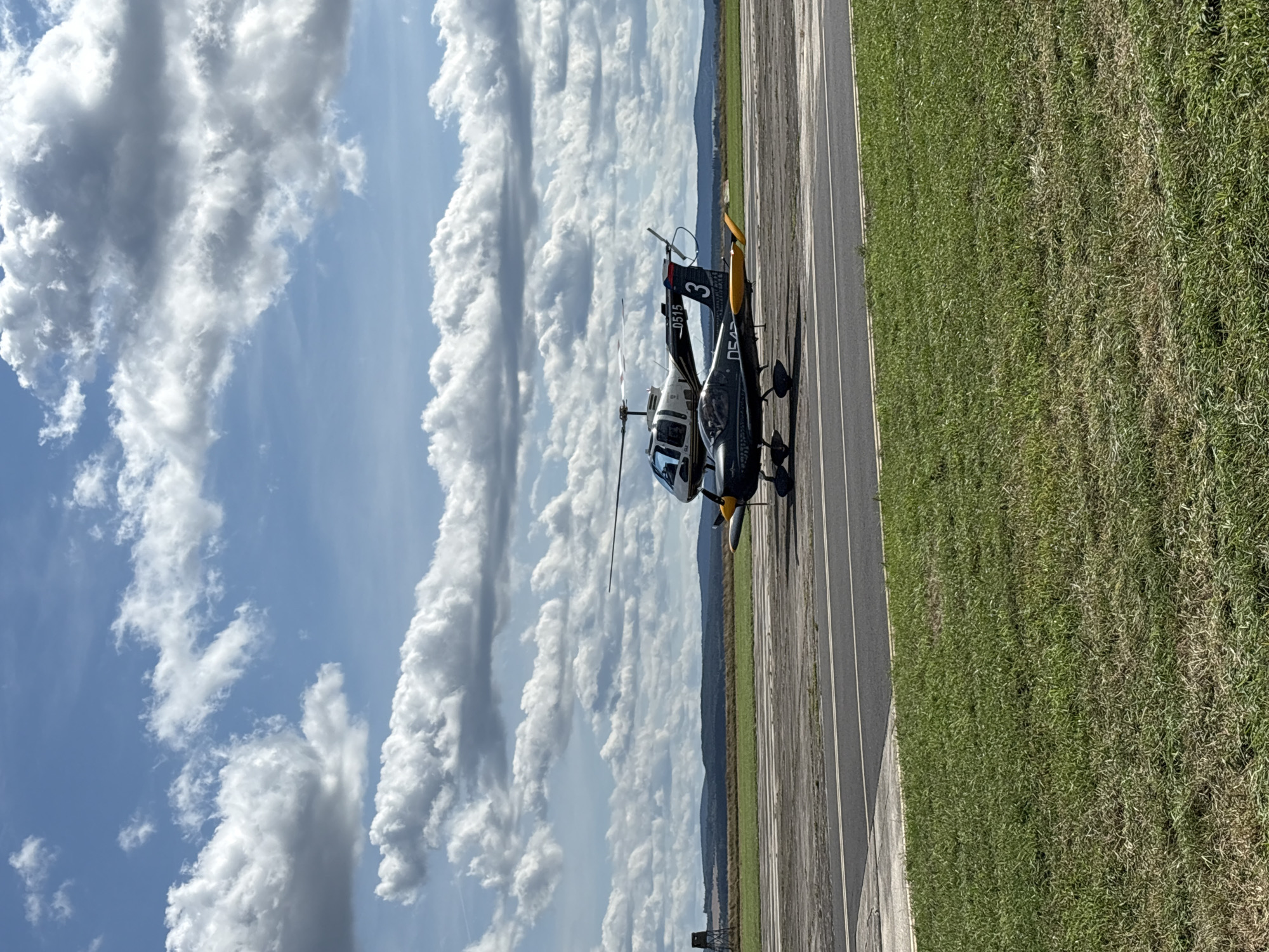 A small airplane is parked on a tarmac next to a helicopter. The airplane is dark colored with yellow wingtips and a number "3" painted on its tail. The helicopter is white with a dark stripe and the registration number "0515" visible. The scene appears to be an airfield. In the background, there are rolling hills and a bright blue sky filled with fluffy white clouds. The grass in the foreground is green and somewhat dry in places. A tall, dark structure, possibly a tower, is visible in the far left background. The location is likely an airfield in or near Cheb, Czechia, given the sky and landscape typical of the region.