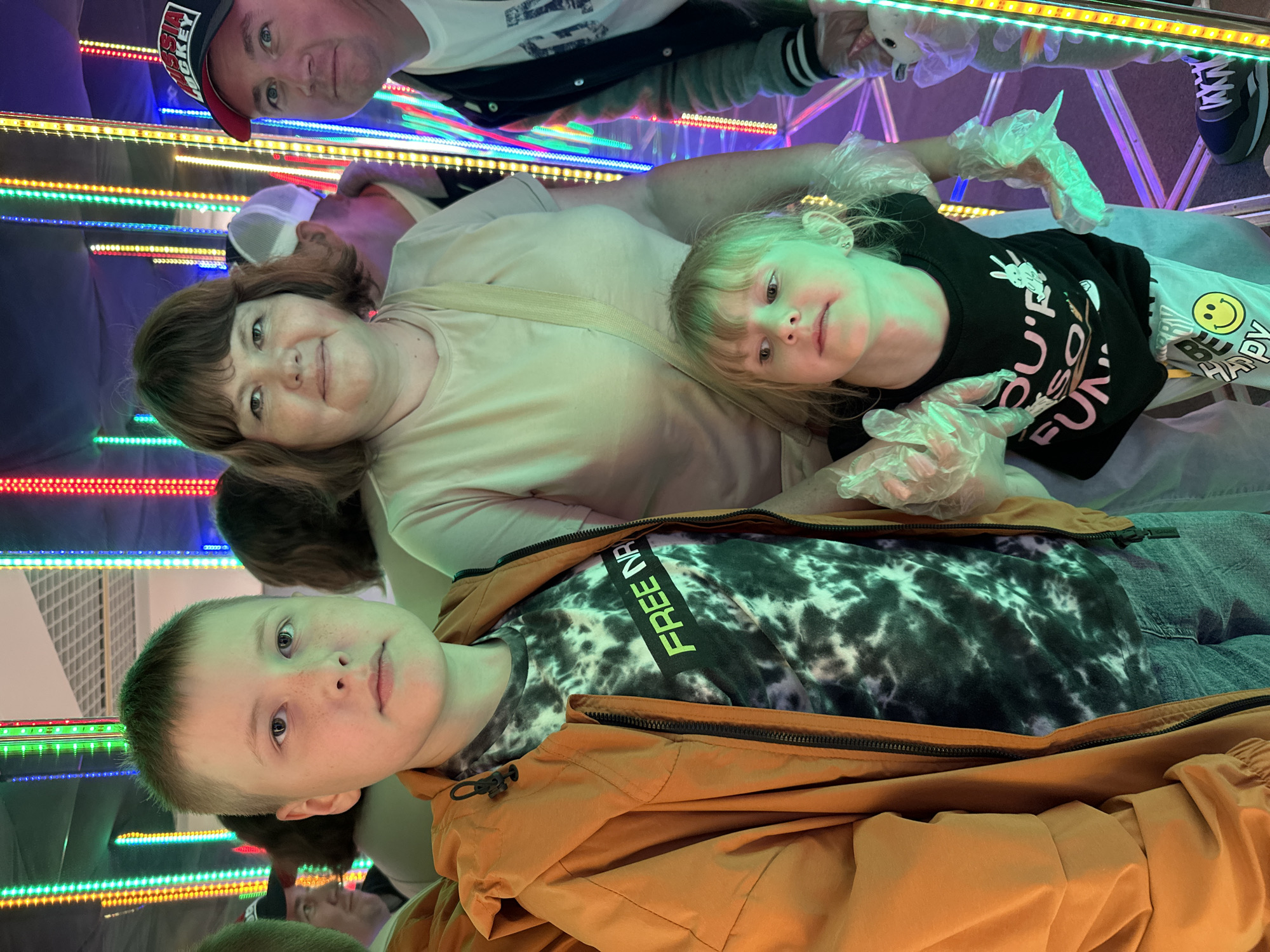 The image shows a family posing for a photo in a brightly lit room with colorful LED lights. In the foreground, a young boy in an orange jacket and a tie-dye shirt stands to the left, looking directly at the camera. To his right, a woman with brown hair and a light-colored t-shirt is positioned behind a young girl wearing a black t-shirt. The girl is looking to the side. To the far right, a man in a black and white jacket and a cap is visible. The environment appears to be an amusement attraction or entertainment venue, possibly in Izhevsk, Russia, given the mention of a "Russia Hockey" cap worn by the man. The overall atmosphere is lively and cheerful, with the colorful lights creating a vibrant backdrop.
