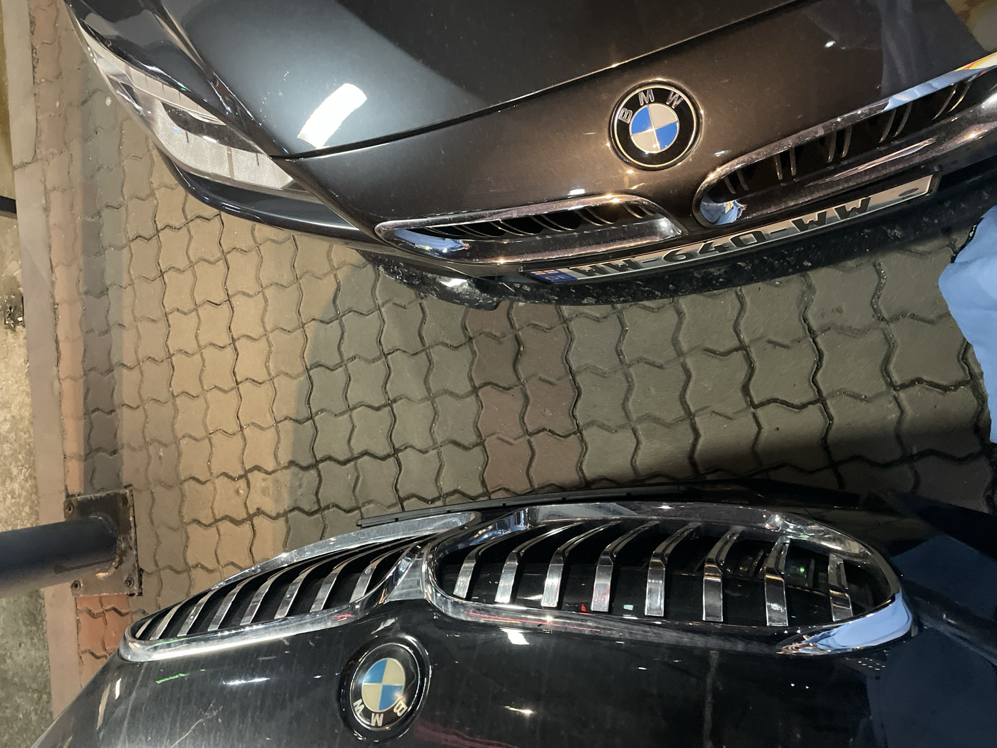 The image depicts the front ends of two BMW cars parked on a herringbone brick pavement. The car at the top of the frame is a dark grey color and its license plate is partially visible. The bottom car is black. The location is possibly Batumi, Georgia. The cars are the main subjects. The pavement is the environment. The action is the cars being parked. There is no interaction depicted. No people are visible.