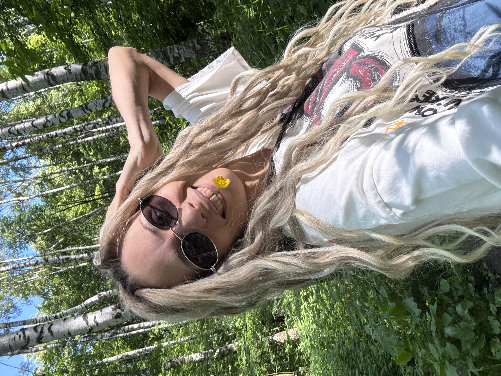 A young woman is depicted in a close-up, upward-angled selfie, standing outdoors in a sunlit forest. She has long, blonde hair styled with a mix of loose waves and several visible dreadlocks or braids. She wears dark, oval-shaped sunglasses and a white t-shirt with a multi-colored graphic design featuring red, black, and blue elements partially visible on the front. A small yellow flower, resembling a buttercup, is held between her teeth on the right side of her mouth, and she is smiling broadly. Her right arm is raised, with her hand positioned near the back of her head. The background consists of numerous birch trees with white and black bark, indicating a forest environment. Lush green foliage and undergrowth cover the ground, and glimpses of a clear blue sky are visible through the tree canopy. The scene takes place in Rusanovo, Russia.