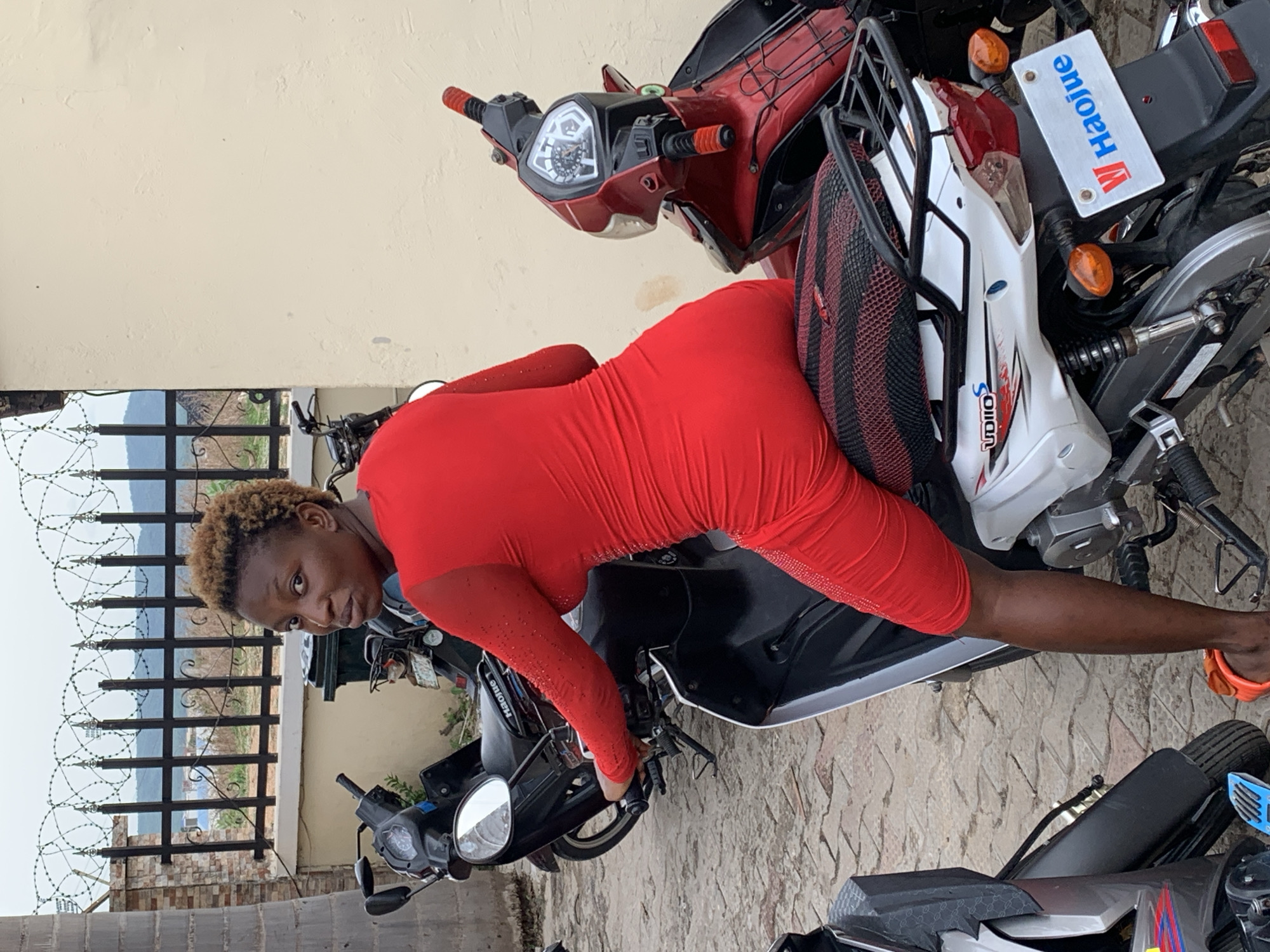A woman with short blonde hair, wearing a form-fitting red dress and orange sandals, is reclining across two parked motorcycles. She appears to be posing for a photo. The motorcycles are of different styles; one is predominantly red and white, while the other is black. A blue and white license plate with the word "Haojue" on it is visible on one of the motorcycles. The background includes a low wall topped with a metal fence and barbed wire, suggesting a possible residential or commercial property in Lokoja, Nigeria. The ground is paved with tiles.