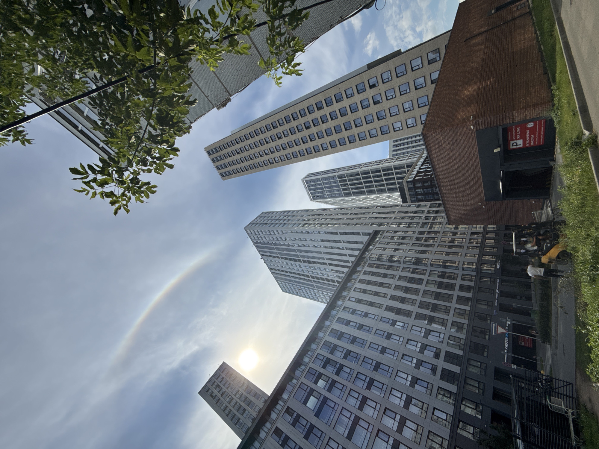 In Moscow, Russia, a low-angle shot captures a cluster of modern, multi-story residential buildings under a cloudy sky. The sun is partially obscured by clouds, creating a bright halo effect, and a faint rainbow is visible in the upper left quadrant of the sky. The buildings, characterized by their glass facades and numerous windows, vary in height and architectural style, with some exhibiting more angular designs. In the foreground, a low-rise brick structure with an entrance to what appears to be an underground parking garage is visible. A yellow forklift with two individuals inside is parked near the brick building, suggesting ongoing work or maintenance. Greenery, including trees and grass, borders the scene, indicating an urban park or landscaped area. The overall impression is one of a contemporary urban environment with a touch of natural phenomena.