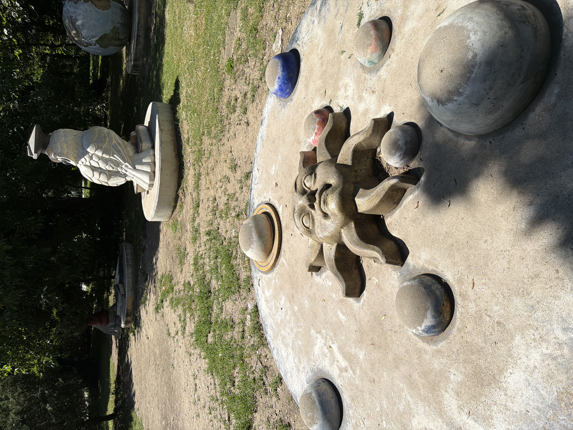 In Pazardzhik, Bulgaria, a solar system-themed sculpture is depicted in an outdoor park setting. The sculpture is set on a concrete circular platform, featuring a prominent sun face at its center, surrounded by seven spherical objects representing planets. These "planets" vary in size and color, with some appearing as grey spheres, one as a blue sphere, and others with hints of red and pink. A brown-toned sun face with closed eyes and a detailed expression is sculpted in the center of the platform.
In the background, a large, white owl statue wearing a graduation cap sits atop a book, also on a circular concrete base. To the right of the owl, a globe sculpture is visible, resting on a similar base. The ground around the sculptures is a mixture of dirt pathways and patches of green grass. Trees with dense foliage provide shade in the background, suggesting a sunny day. The overall environment appears to be a public park or a sculptural garden.