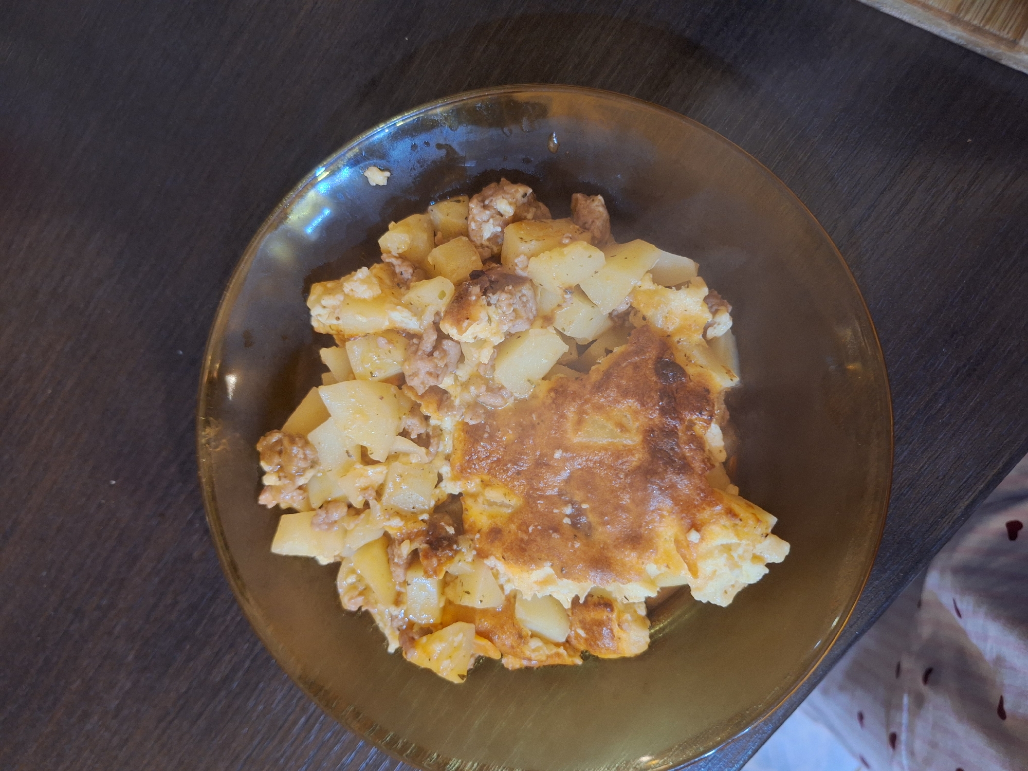 The image shows a plate of food on a dark brown wooden surface. The food appears to be a potato and meat casserole with a browned crust. Steam or vapor is rising from the dish, suggesting it is freshly cooked. The plate is golden or amber in color. Part of a patterned cloth is visible in the lower right corner. The location is not discernible from the image.