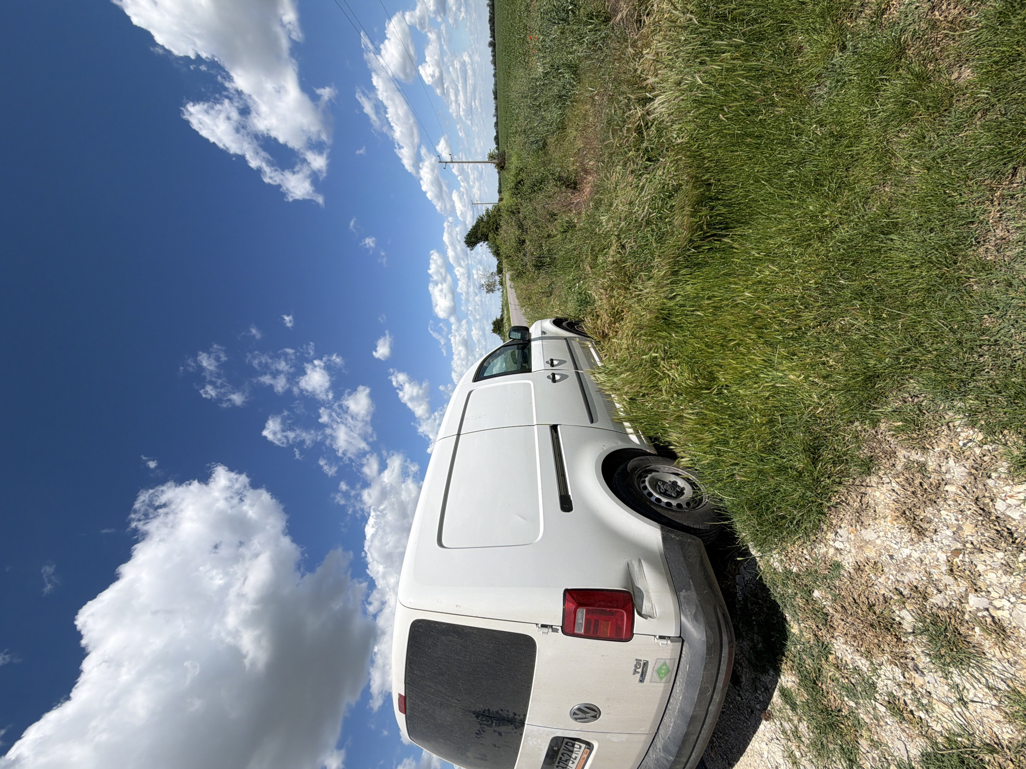 A white Volkswagen Caddy van is parked on a dirt road next to a grassy field. The sky is blue with scattered white clouds. Utility poles are visible in the distance. The photograph was likely taken outdoors in a rural area near Dve Mogili, Bulgaria.