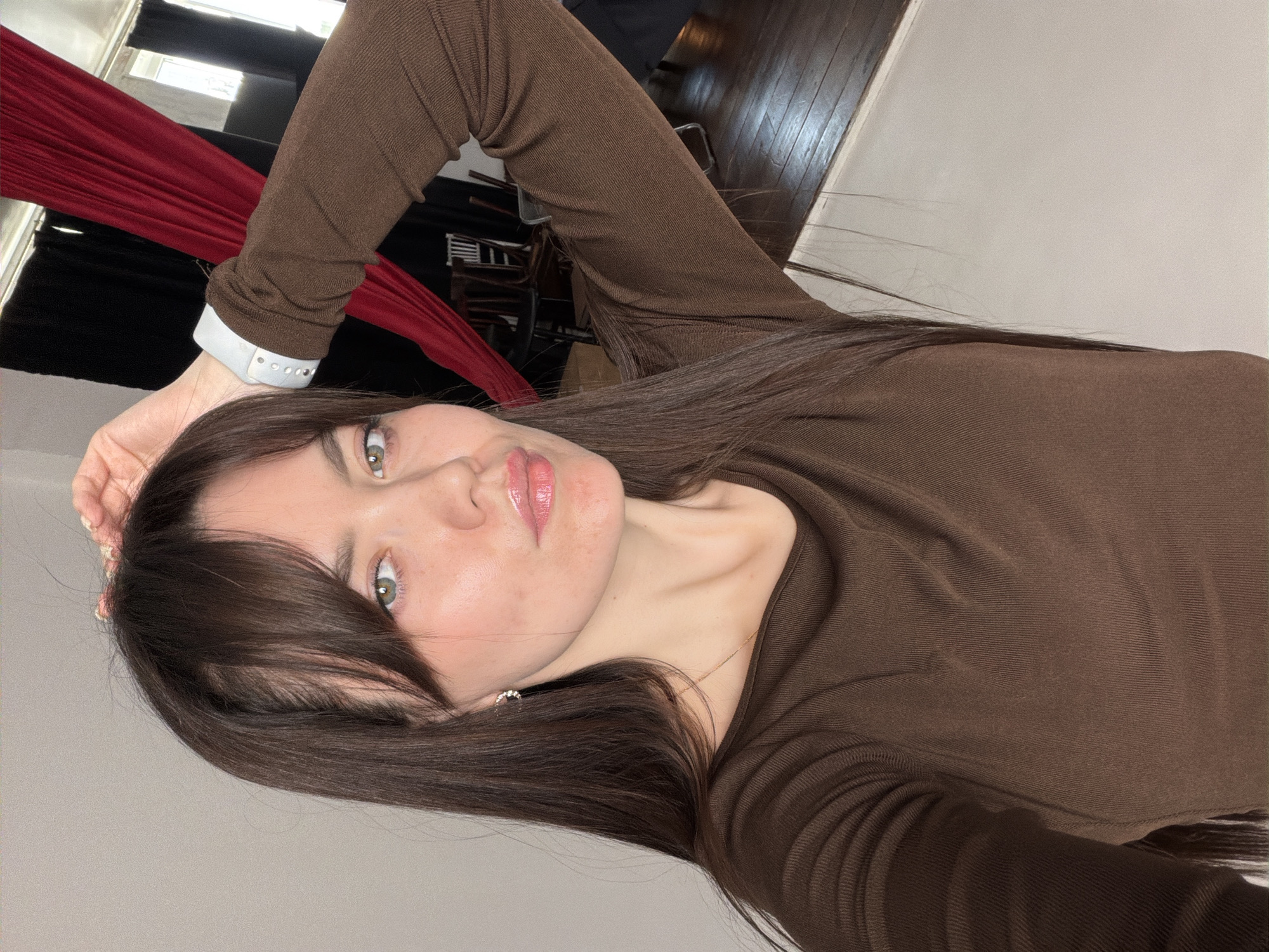 A woman with shoulder-length dark hair, parted on the side, is lying down and taking a selfie indoors. She is wearing a long-sleeved brown top. Her left arm is raised, with a white device around her wrist. Behind her are visible red fabric and dark curtains, and a glimpse of a hardwood floor. The location is potentially in Chaikovsky, Russia.