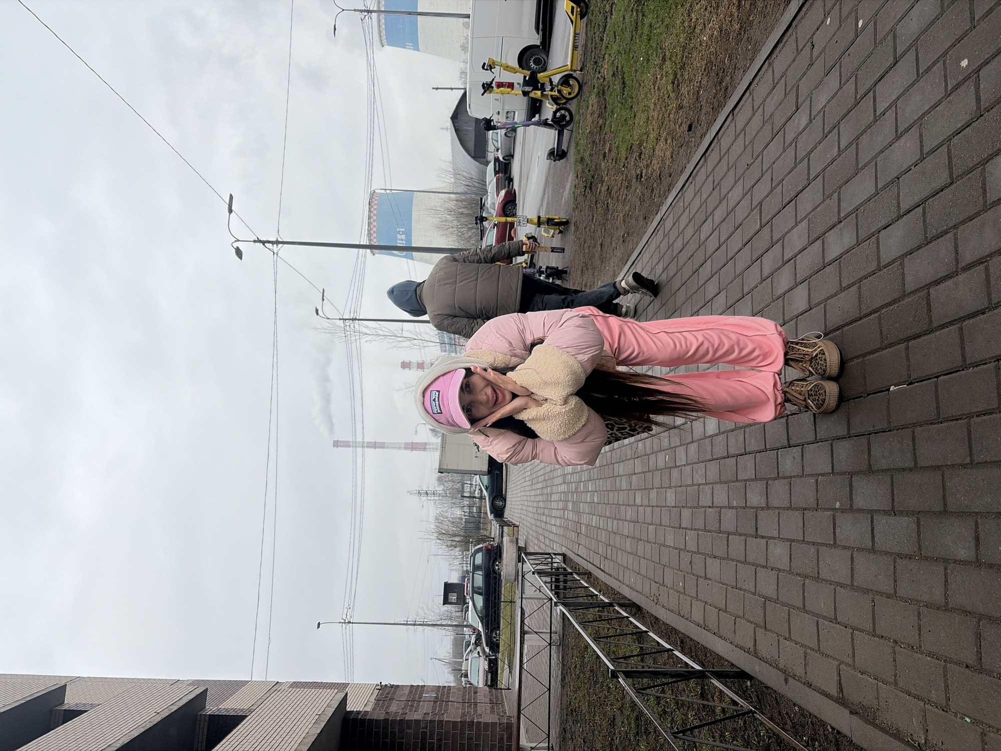 The image shows a woman in a pink tracksuit posing on a brick sidewalk, her hands framing her face. She is wearing a pink beanie and leopard-print shoes. Behind her, a man is interacting with electric scooters. In the background are parked cars, a building, and a smokestack. Based on the smokestack and overall environment, the location may be in the city of Saint Petersburg, Russia.