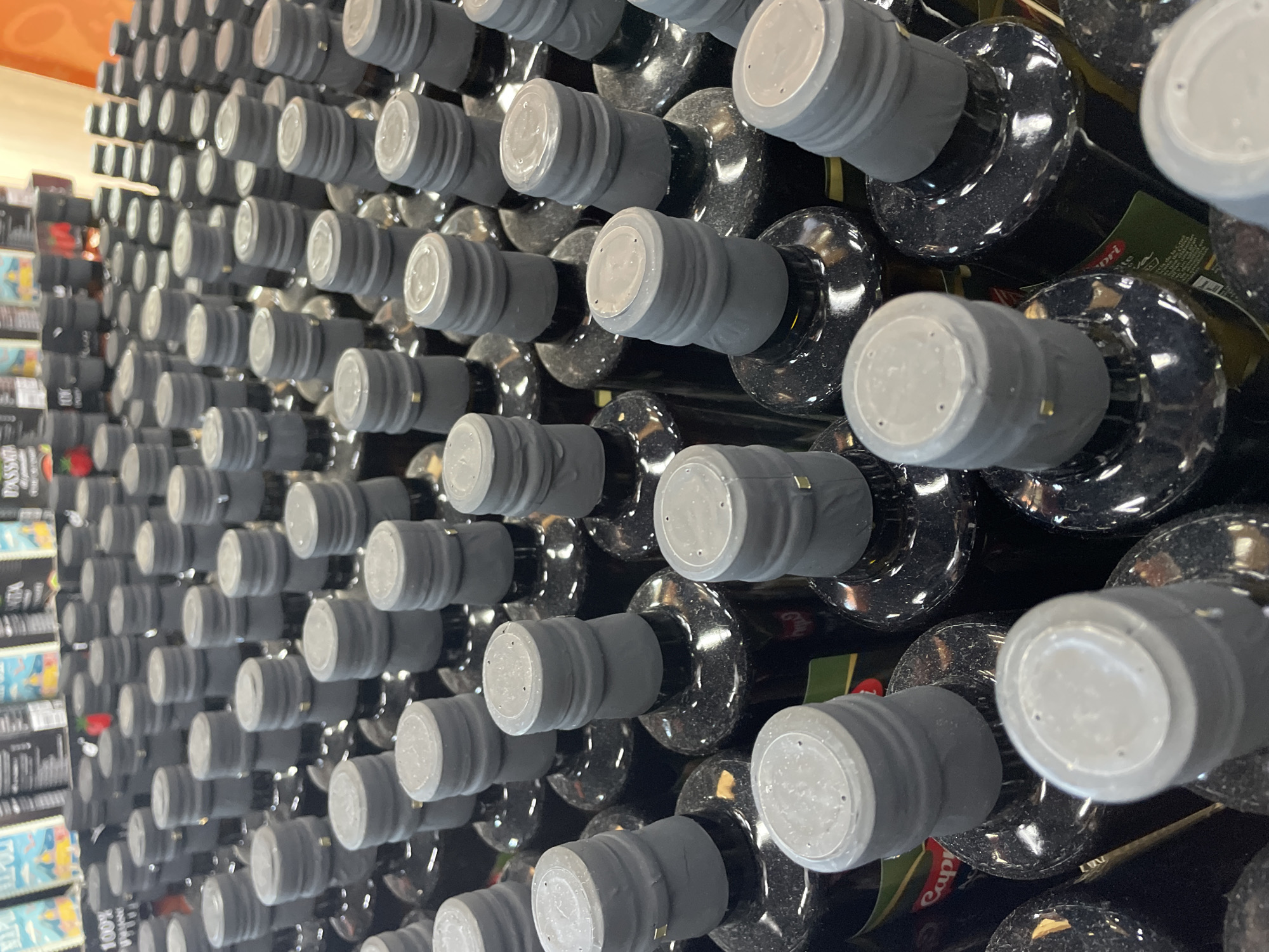 The image shows a close-up, overhead view of numerous dark glass bottles with grey caps arranged in rows. The bottles appear to be olive oil bottles, with some labels visible in green and red that suggest a brand name such as "Capri." The caps are ribbed and have a metallic seal at the bottom. The bottles are dusty, indicating they might be on display in a store. The background is out of focus but suggests shelving with more products. There are no people or animals visible in the image. The lighting is bright and appears to be artificial, reflecting off the glass bottles. The scene is likely from a supermarket or specialty food store.