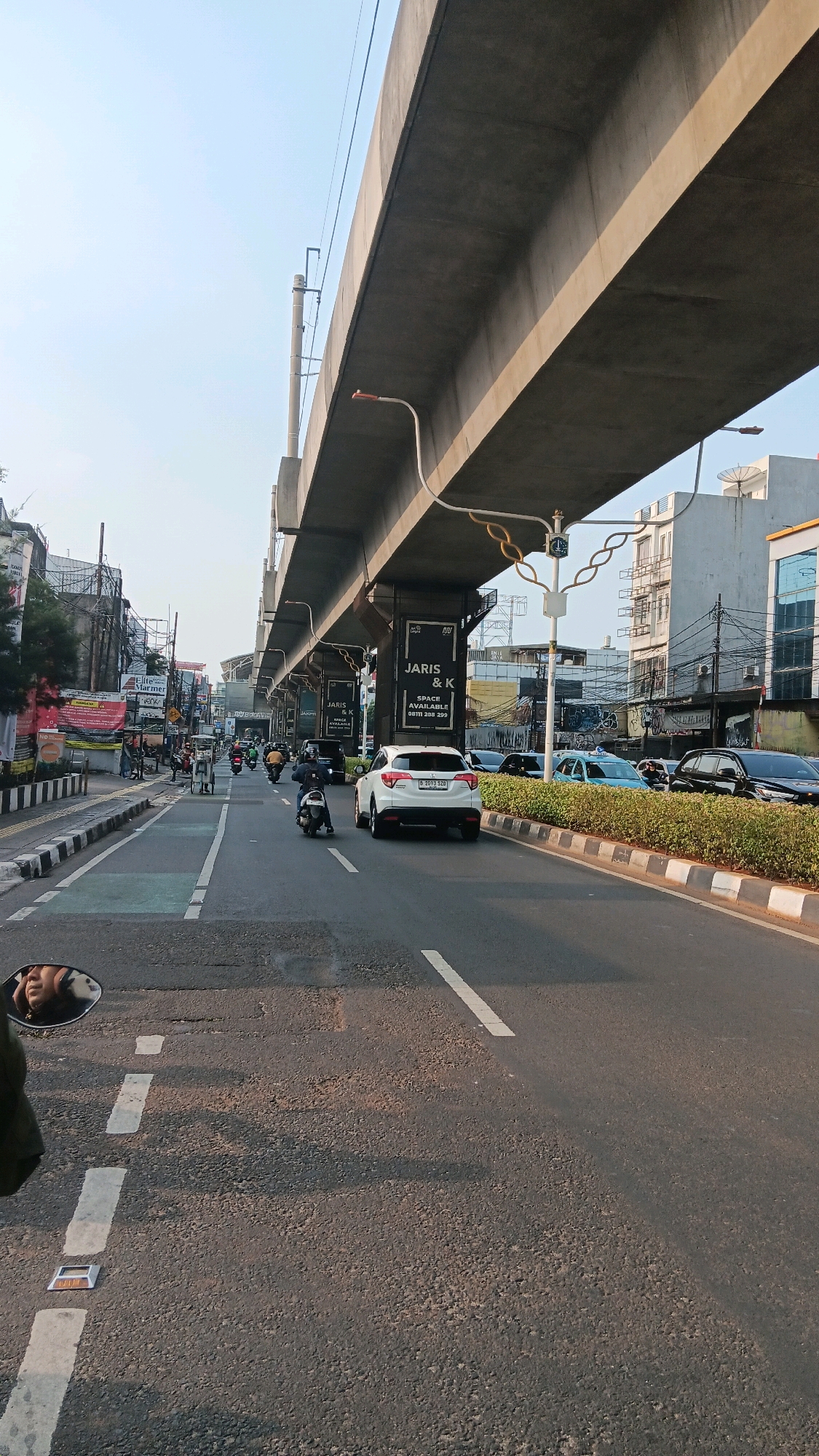 The image depicts a street scene in South Jakarta, Indonesia, likely during the daytime. A multi-level overpass dominates the upper portion of the frame, casting shadows on the road below. The road itself is asphalt with white lane markings and is populated by several vehicles, including motorcycles and cars. A white Honda SUV is prominently featured in the right lane, its license plate visible. On the left side of the road, there's a sidewalk with some vegetation and buildings in the background. Banners are hanging from the overpass. In the foreground, a motorcycle's side mirror reflects a person's face, suggesting the photo was taken from the perspective of a motorcyclist. The overall environment appears to be an urban setting with moderate traffic.