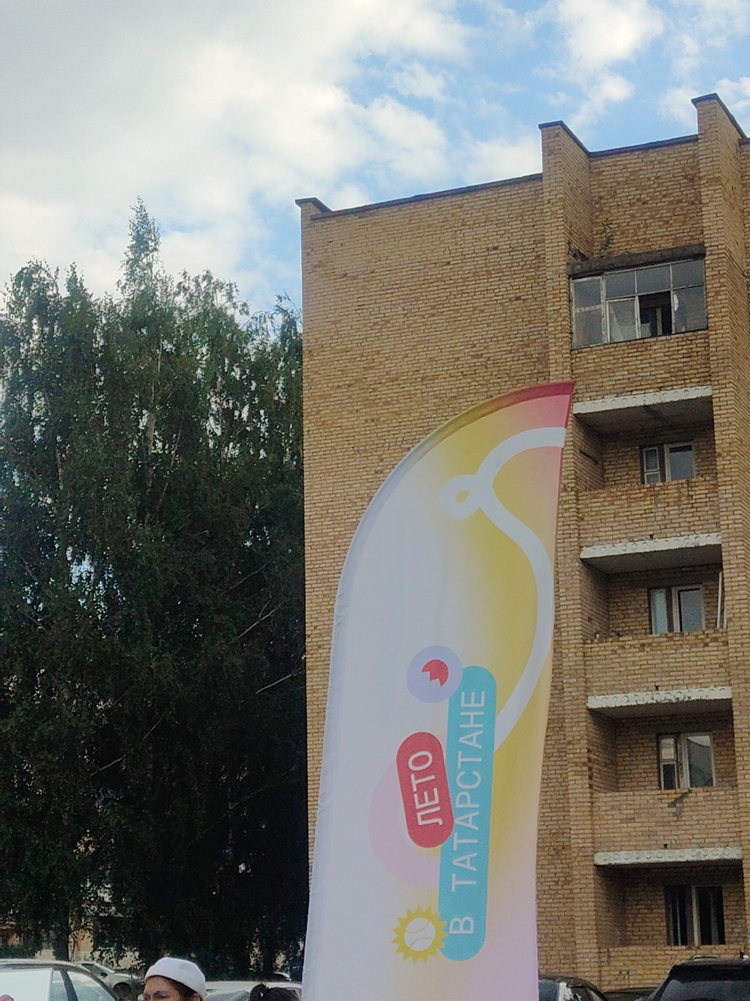 The image shows a partial view of a light brown brick building with several visible windows and balconies. To the left of the building is a large, dark green tree with dense foliage. In the foreground, a large, curved banner with colorful graphics and text is visible. The text on the banner appears to be in Russian, with "ЛЕТО" (Summer) and "В ТАТАРСТАНЕ" (In Tatarstan) prominently displayed. Below "ЛЕТО" is a stylized icon of a sun. At the bottom left of the image, part of a person's head wearing a white cap is visible. Several cars are parked in the background, obscured by the banner and tree. The sky is partly cloudy. The location is specified as Mendeleyevsk, Russia.