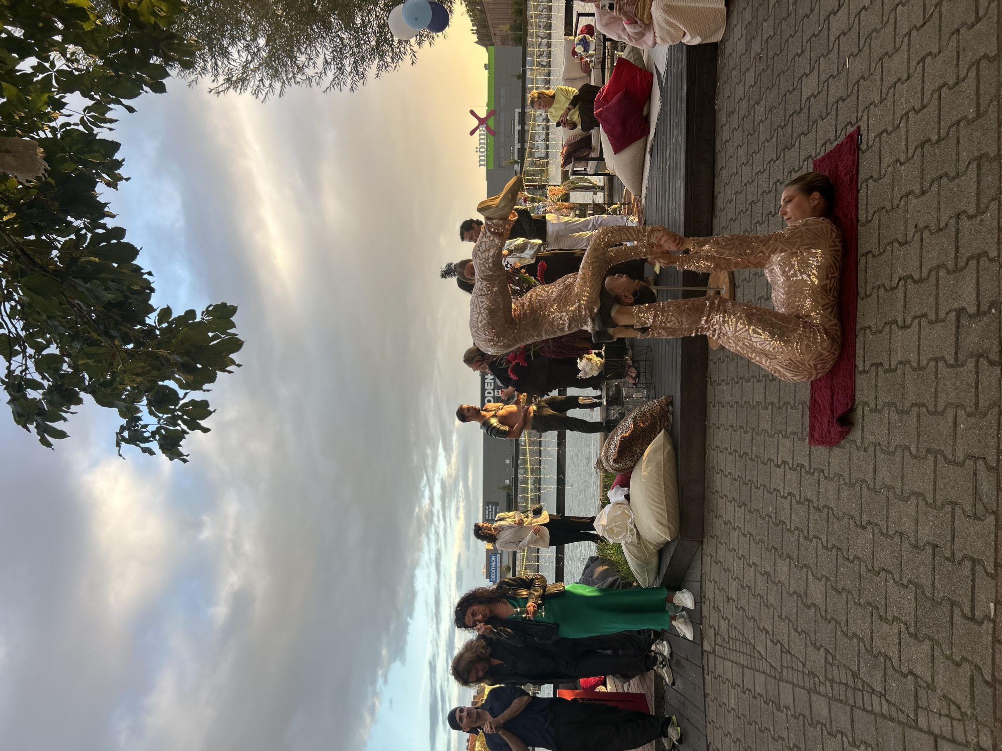 The image depicts an outdoor event on a paved walkway near a body of water. In the foreground, two acrobats are performing, one lying on their back with their legs raised, and the other balanced on the feet of the person on the ground. Both are wearing sequined outfits.
In the middle ground, a group of people are gathered, some standing and observing, while others are seated on cushions. They appear to be attending a performance or social gathering. Behind them, a railing separates the walkway from the water, and across the water, a large building with the "MÖMAX" logo is visible. The sky is overcast with hints of sunset colors. The overall atmosphere suggests a relaxed, evening event. This scene is likely taking place in Berlin, Germany, given the presence of the MÖMAX store, which is a well-known furniture retailer with locations in the city.