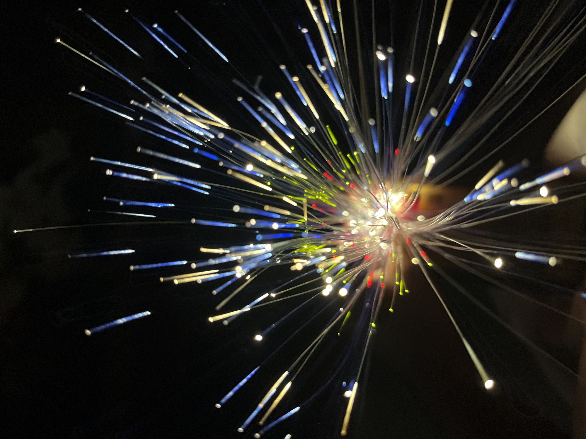 The image shows a fiber optic lamp against a dark background. The fibers emanate from a central point and are lit in various colors, including white, blue, red, and green. The central point is brightly illuminated. The scene appears to be indoors. No people are visible. There is no information in the image to indicate a location in Dhaka, Bangladesh.